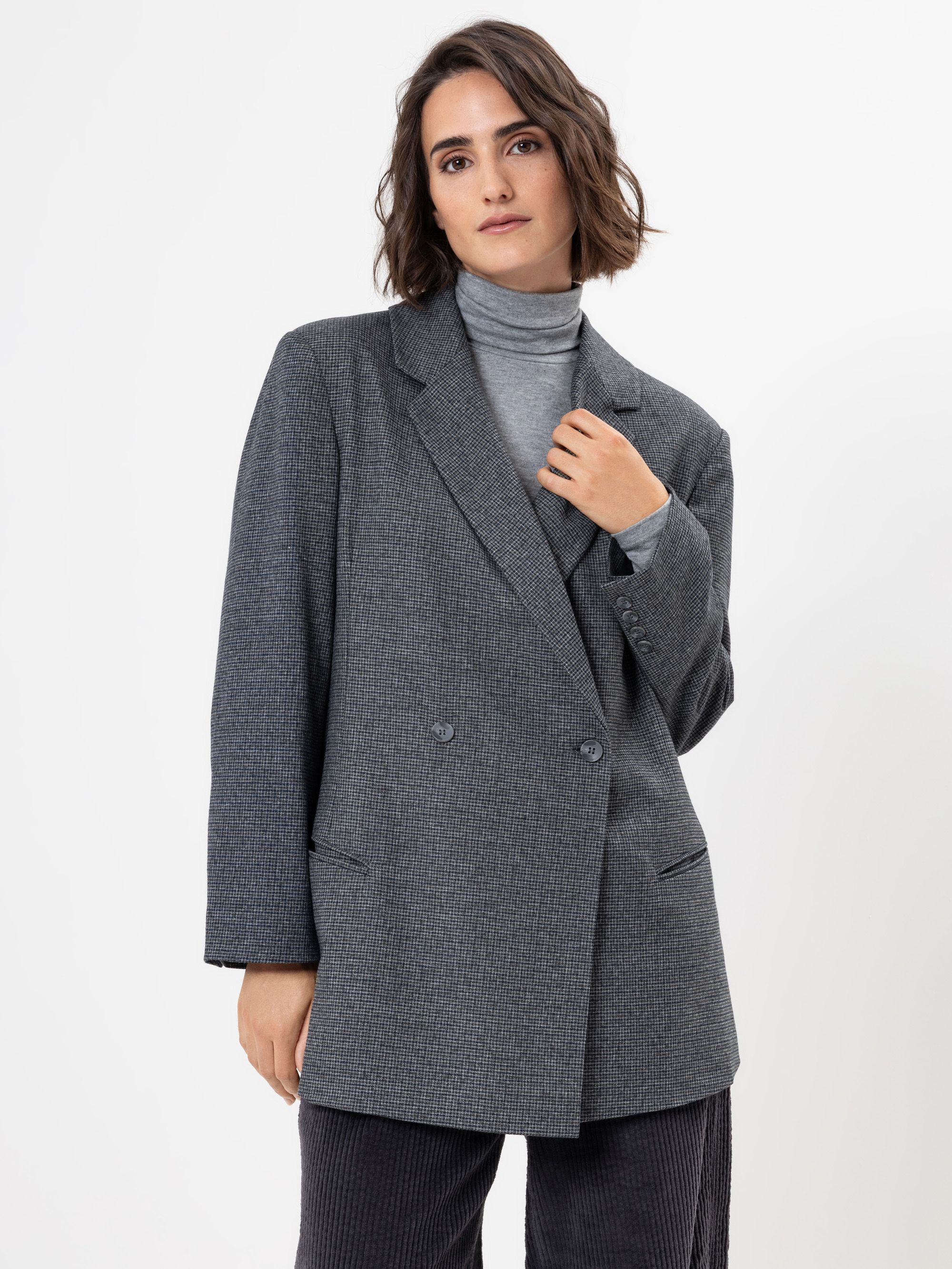 Eine Frau mit kurzen braunen Haaren, die einen grauen doppelreihigen Blazer über einem hellgrauen Rollkragenpullover trägt, steht vor einem schlichten weißen Hintergrund und schaut selbstbewusst in die Kamera.