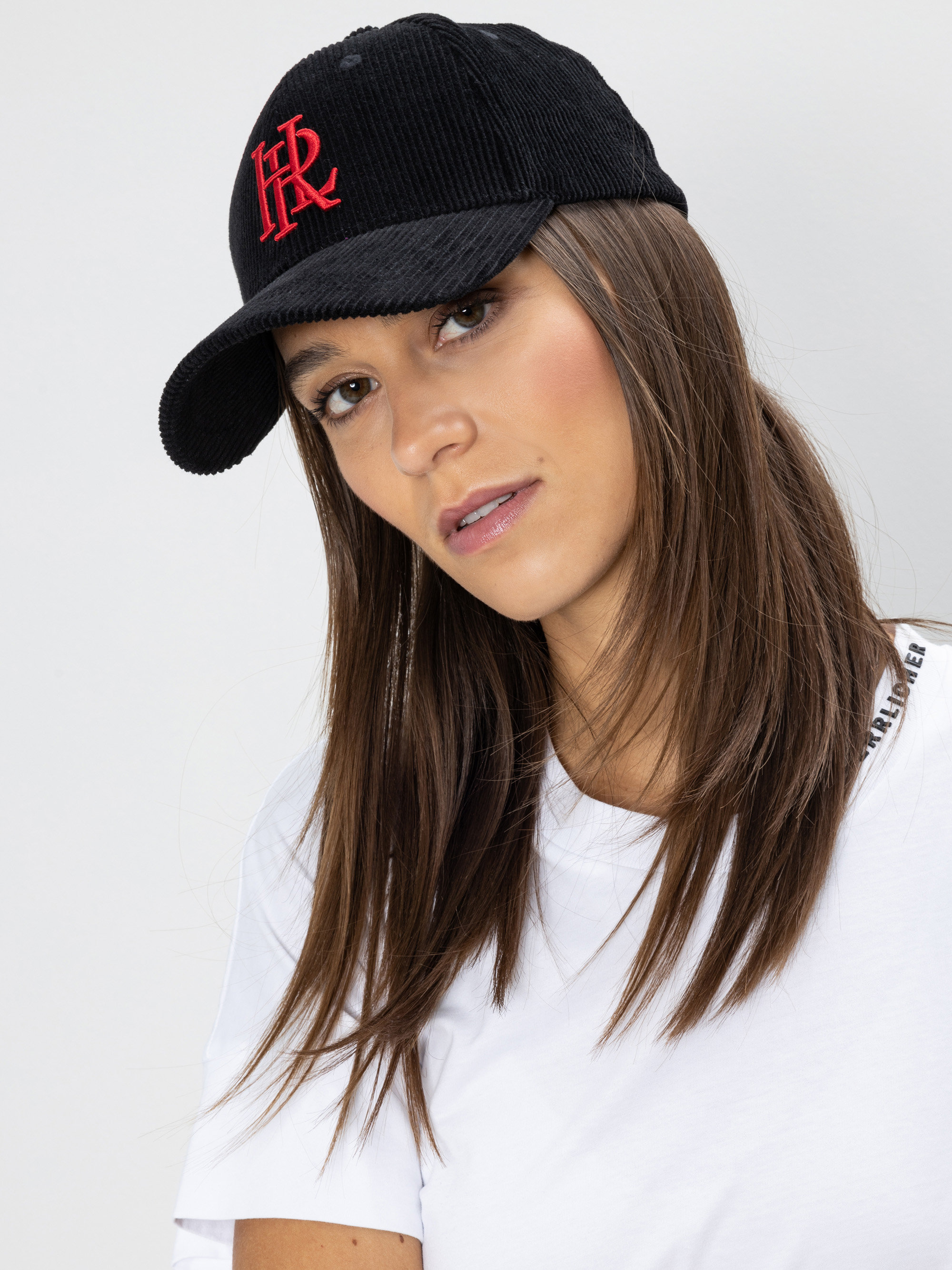 Eine Frau mit langen braunen Haaren trägt eine schwarze Mütze mit einem roten aufgestickten Logo und ein weißes T-Shirt und blickt leicht zur Seite vor einem schlichten hellen Hintergrund.