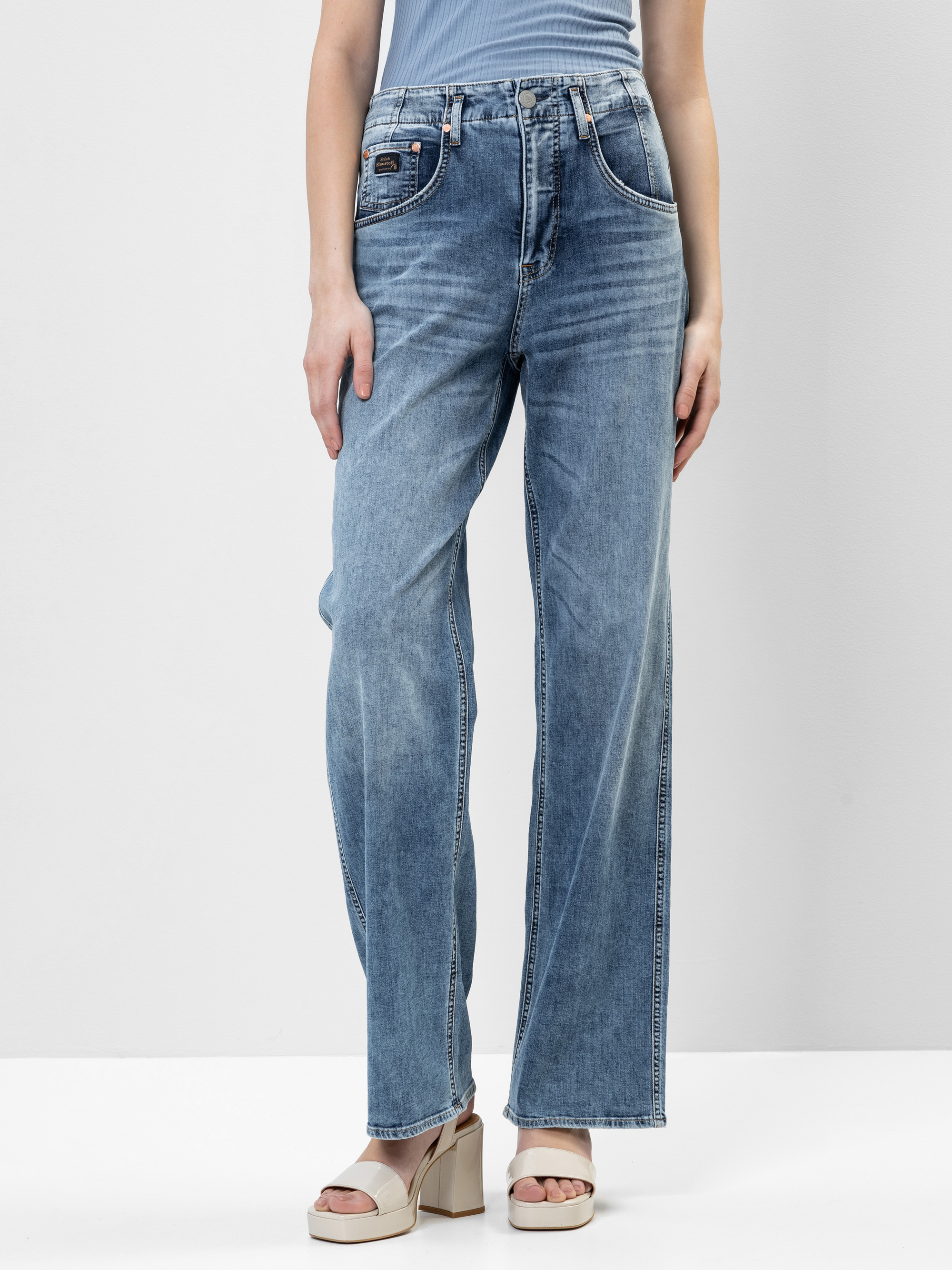 Eine Person, die eine hochtaillierte, locker sitzende blaue Jeans mit leichtem Fading, ein hellblaues geripptes Tanktop und beigefarbene Plateausandalen trägt, steht vor einem schlichten hellen Hintergrund.