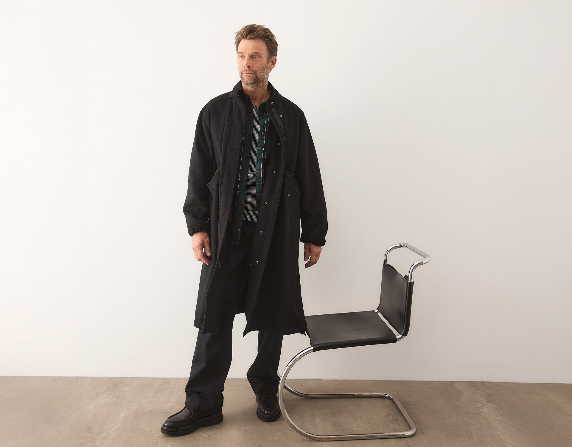 Ein Mann, der einen langen schwarzen Mantel über einem karierten Hemd und einer dunklen Hose trägt, steht neben einem modernen schwarz-verchromten Stuhl in einem minimalistischen, hellen Raum.
