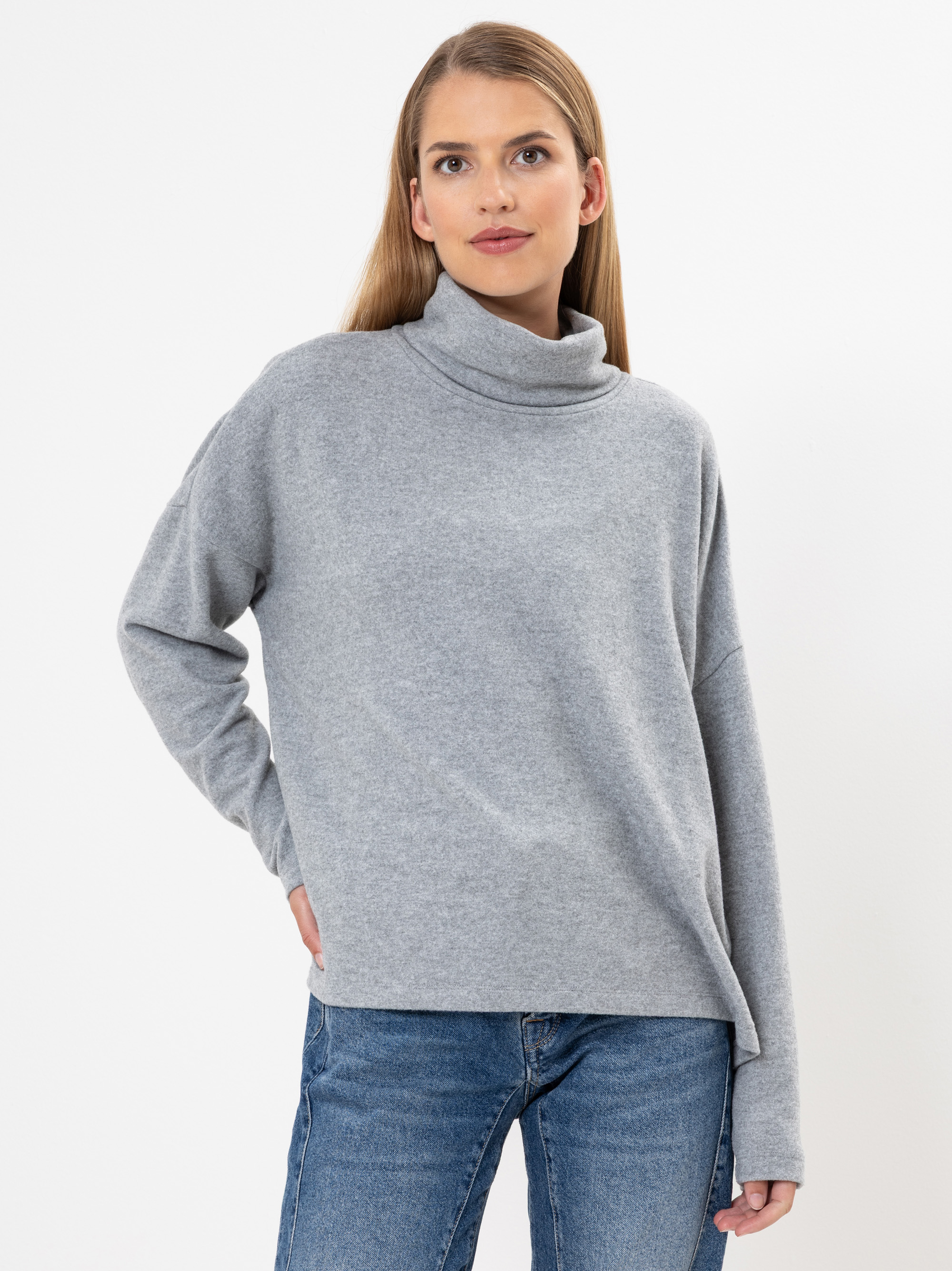 Eine Frau, die einen hellgrauen Rollkragenpullover und blaue Jeans trägt, steht vor einem schlichten weißen Hintergrund, eine Hand in die Hüfte gestützt und mit neutralem Gesichtsausdruck.
