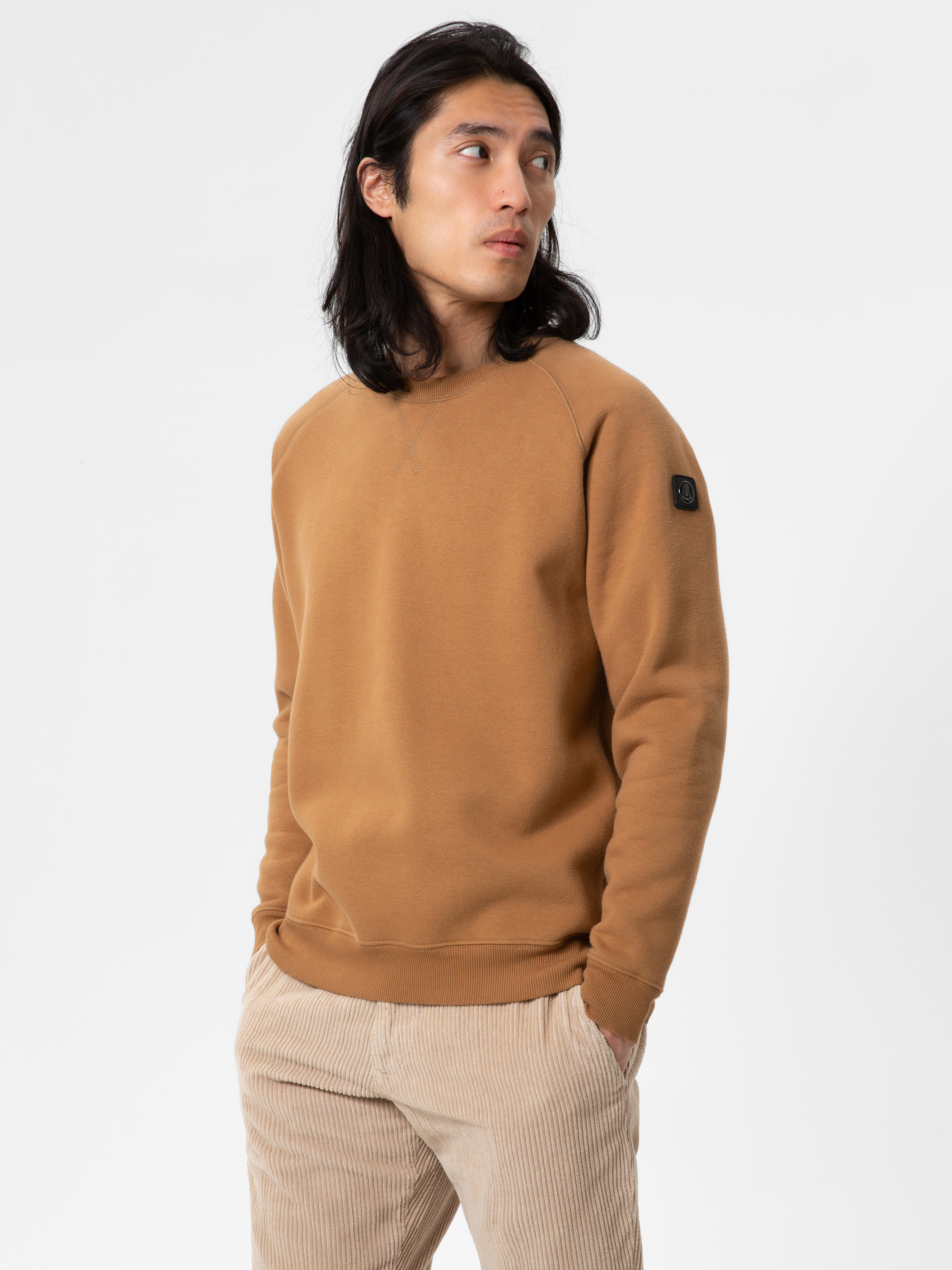 Eine Person mit langen dunklen Haaren, die ein hellbraunes Sweatshirt und eine beigefarbene Cordhose trägt, steht mit den Händen in den Taschen und blickt zur Seite vor einem schlichten weißen Hintergrund.