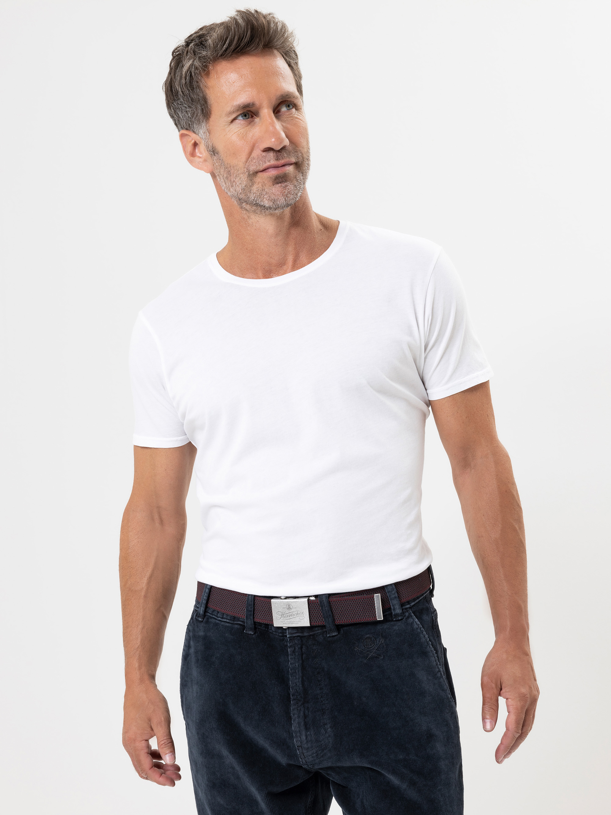 Ein Mann mit kurzen braunen Haaren, etwas grau, und einem gestutzten Bart trägt ein schlichtes weißes T-Shirt und eine dunkle Hose. Er steht vor einem schlichten weißen Hintergrund und blickt mit entspanntem Gesichtsausdruck leicht zur Seite.