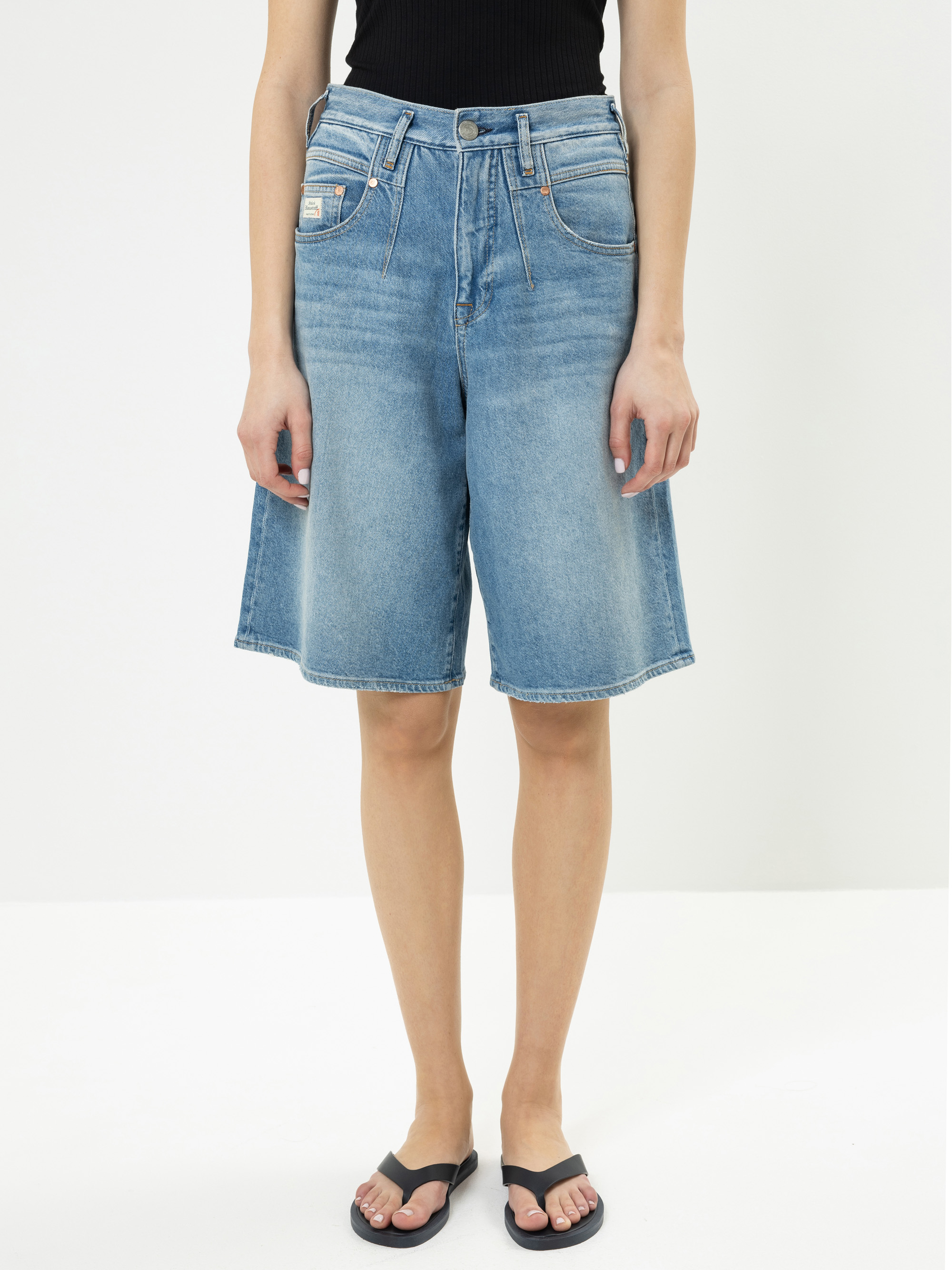 Eine Person, die ein schwarzes ärmelloses Oberteil, knielange blaue Jeansshorts und schwarze Flip-Flops trägt, steht vor einem einfarbig weißen Hintergrund. Das Foto zeigt das Outfit von den Schultern abwärts.