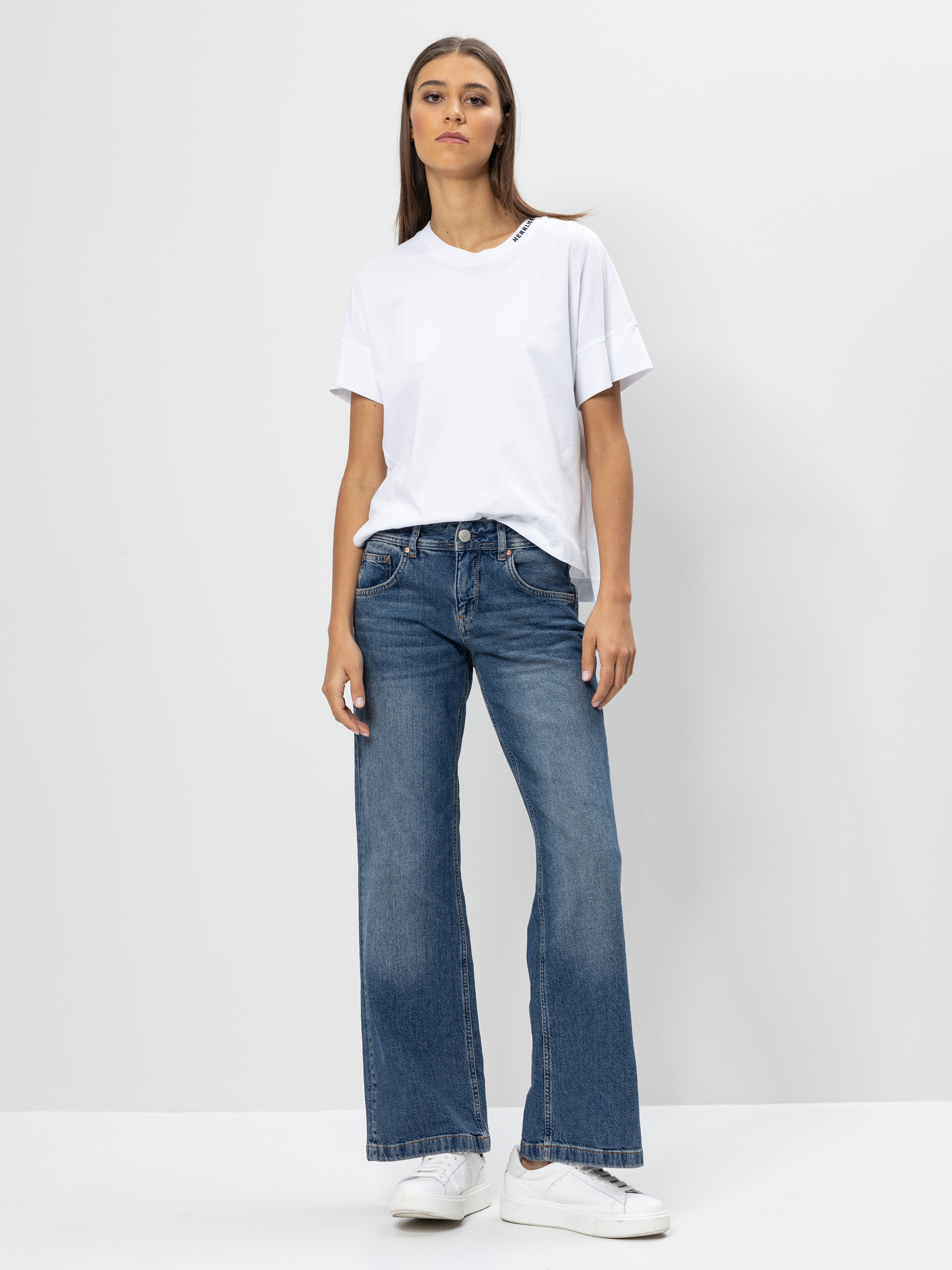 Eine Frau steht vor einem schlichten weißen Hintergrund, trägt ein weißes T-Shirt, eine blaue Jeans mit weitem Bein und weiße Turnschuhe. Sie hat die Arme entspannt an den Seiten und einen neutralen Gesichtsausdruck.