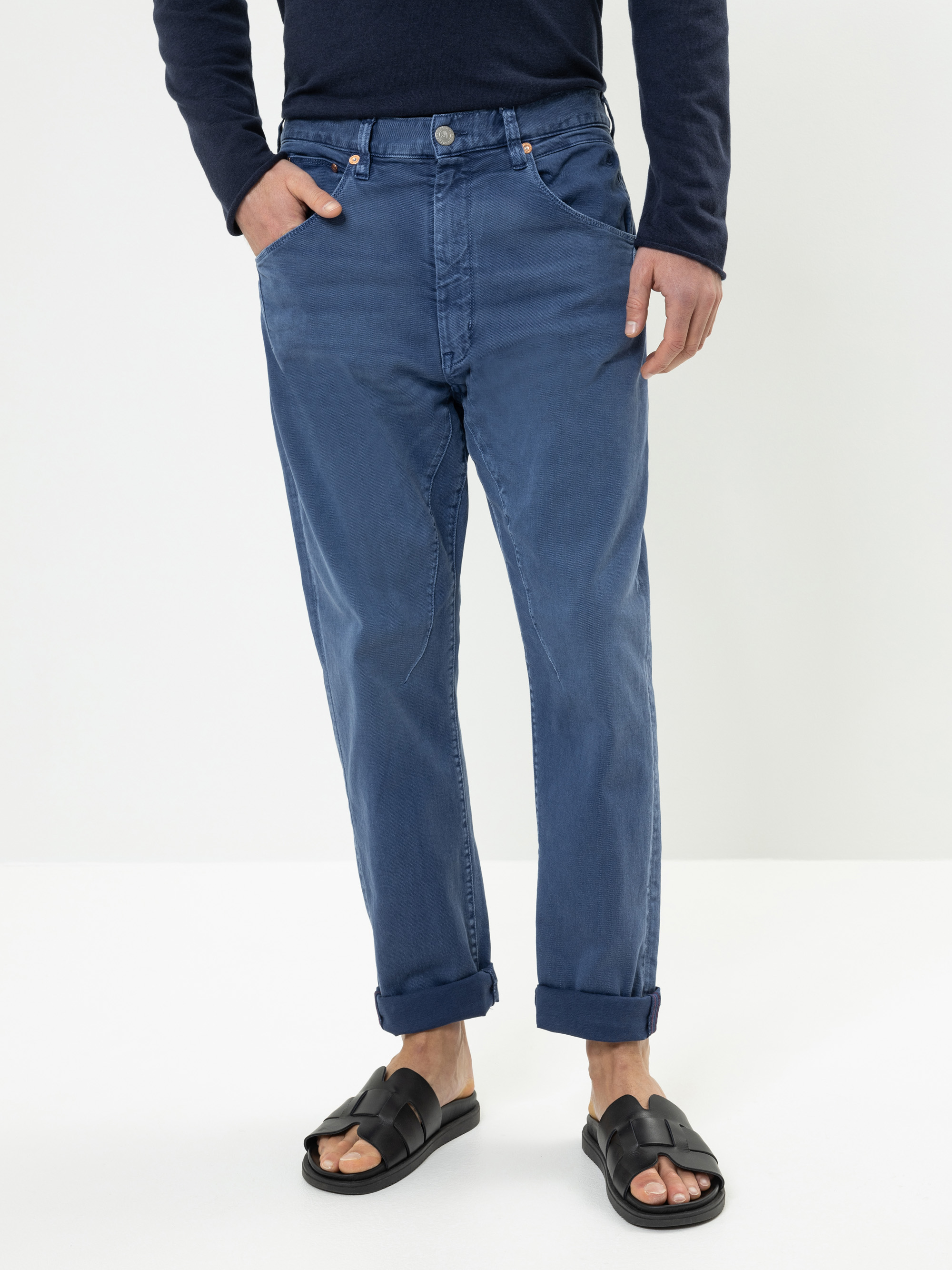 Eine Person, die eine blaue, locker sitzende Jeans mit Manschetten, ein marineblaues, langärmeliges Hemd und schwarze Sandalen trägt, steht mit den Händen in den Taschen vor einem schlichten weißen Hintergrund.