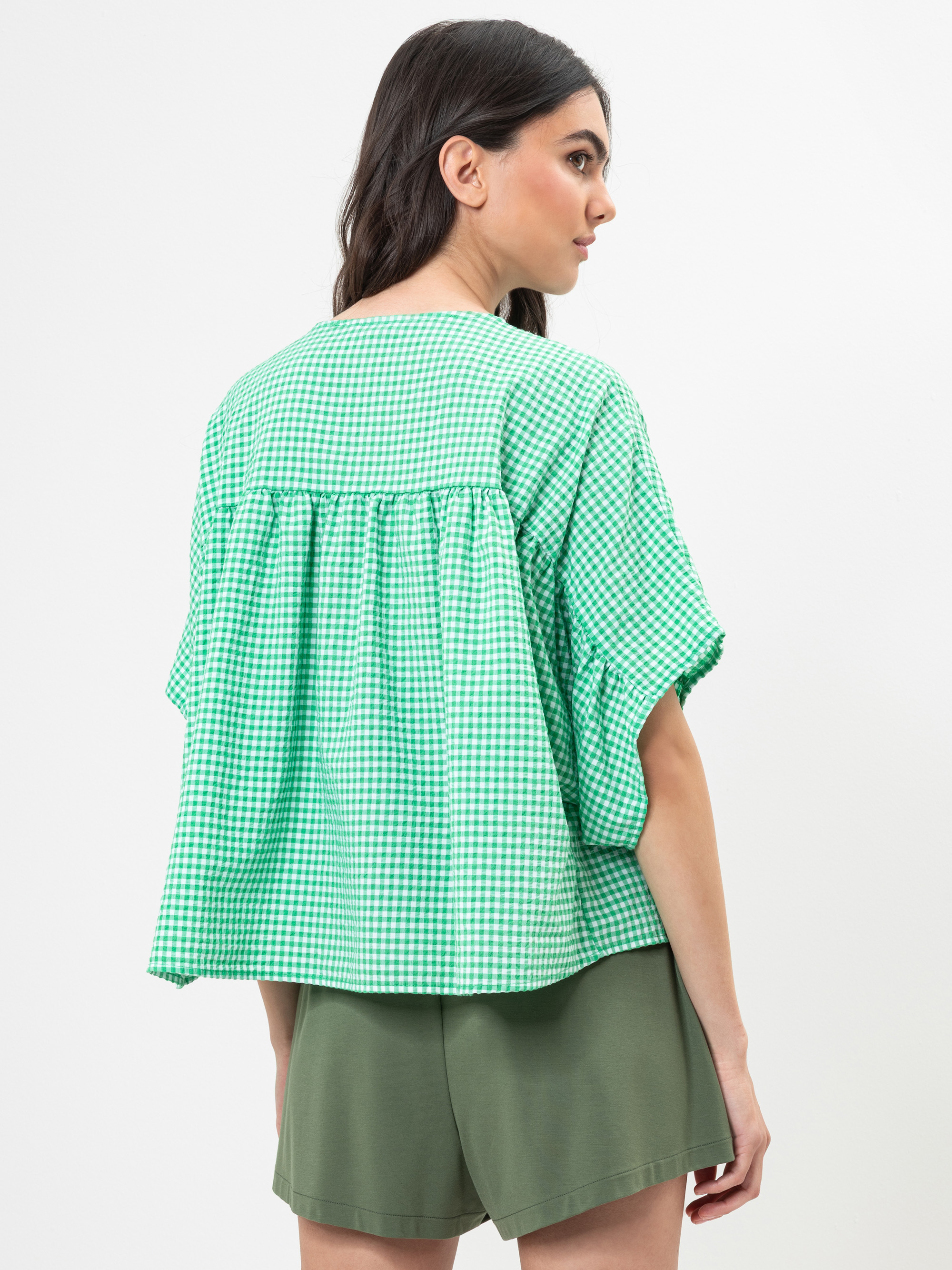Eine Frau mit langen dunklen Haaren trägt eine locker sitzende, grün-weiß karierte Bluse mit kurzen Ärmeln und olivgrüne Shorts. Sie steht vor einem schlichten weißen Hintergrund und blickt leicht zur Seite.