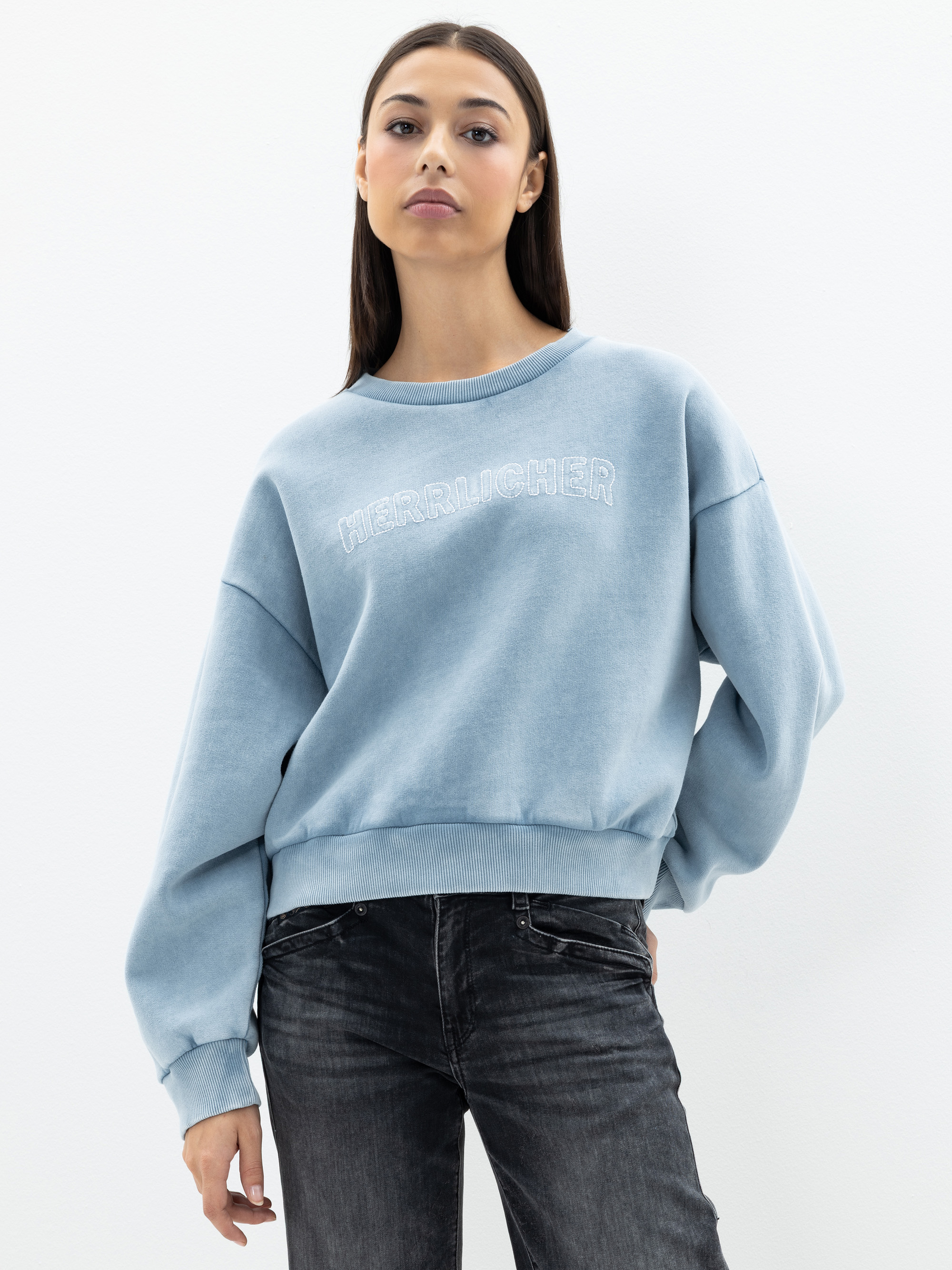 Eine Frau mit glattem braunem Haar trägt ein hellblaues Sweatshirt mit weißer Aufschrift und dunkle Jeans. Sie steht vor einem schlichten weißen Hintergrund und wirkt entspannt.