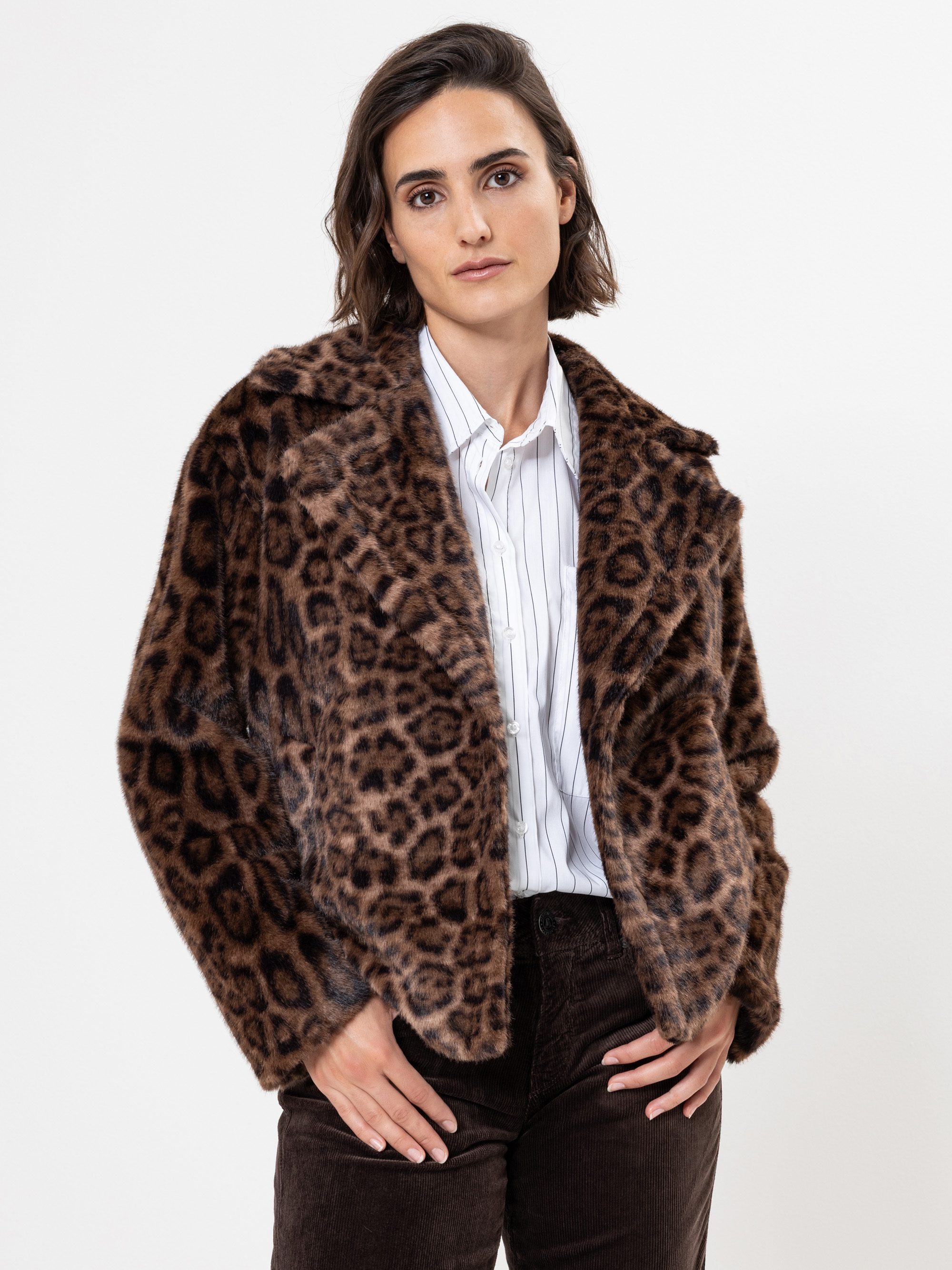 Eine Person mit kurzen braunen Haaren trägt ein weißes gestreiftes Hemd, eine braune Jacke aus Kunstpelz mit Leopardenmuster und eine dunkelbraune Hose und steht vor einem schlichten weißen Hintergrund.
