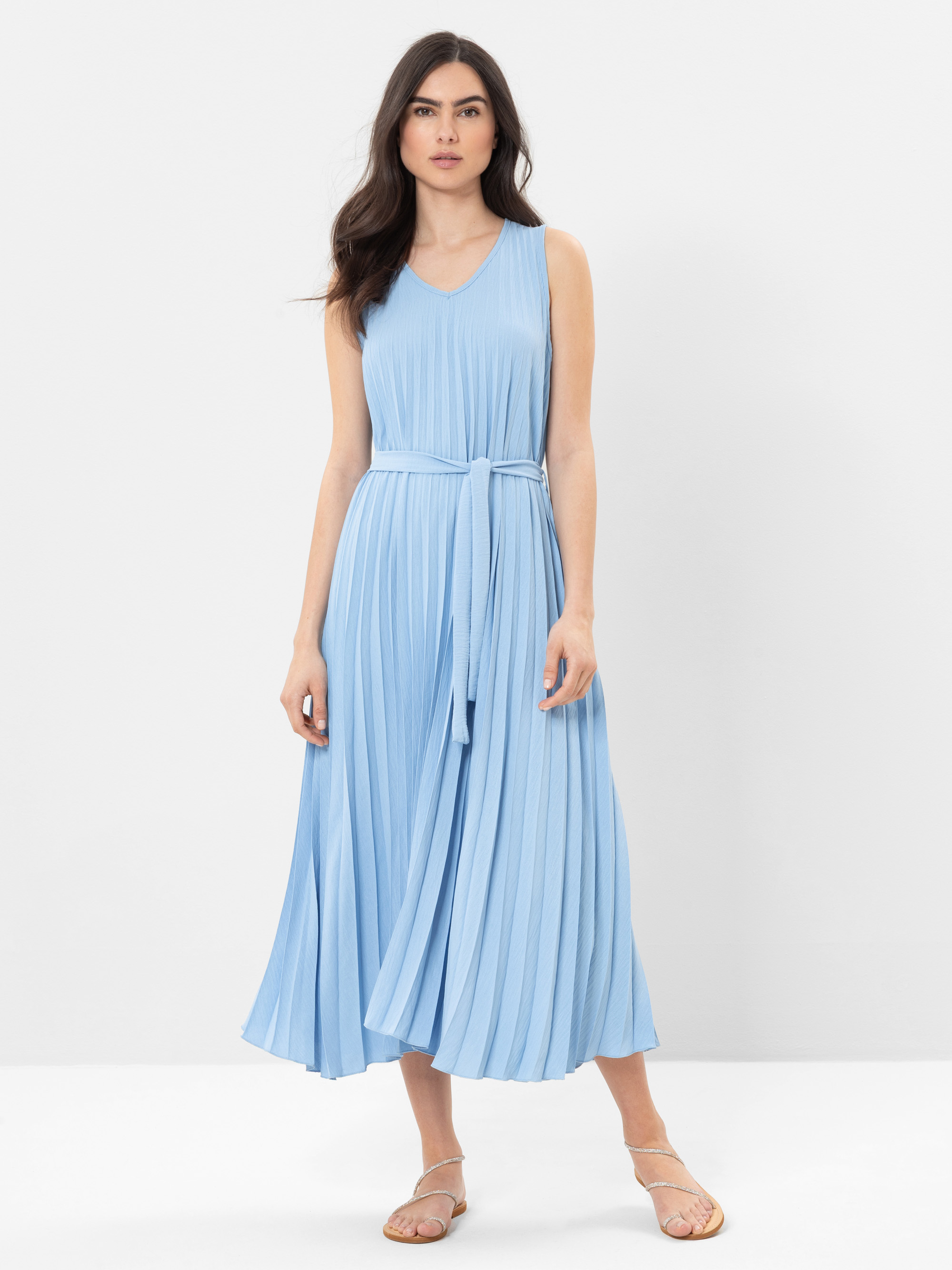 Eine Frau in einem ärmellosen, hellblauen, plissierten Midikleid mit passendem Gürtel und beigen Sandalen steht vor einem schlichten weißen Hintergrund.