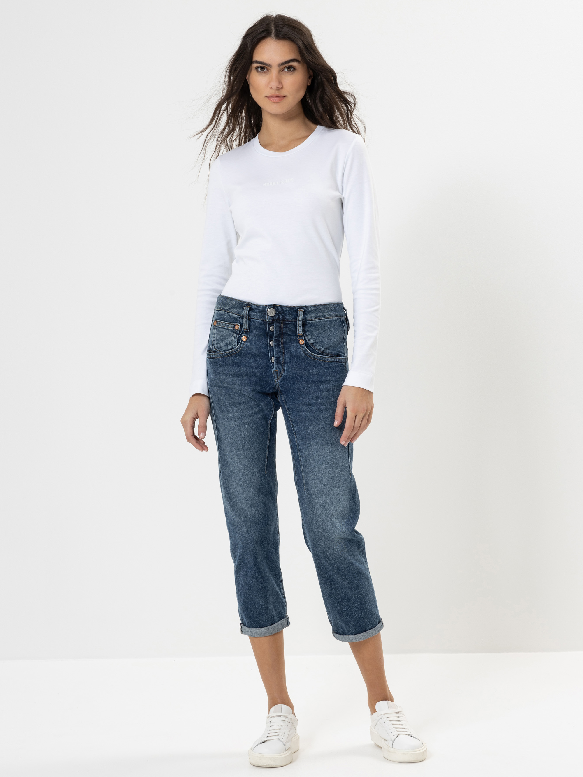 Eine Frau steht vor einem schlichten weißen Hintergrund, trägt ein langärmeliges weißes Hemd, eine blaue Jeans mit aufgerollten Manschetten und sichtbaren Knöpfen und weiße Turnschuhe. Sie blickt geradeaus und hat die Arme entspannt an der Seite.