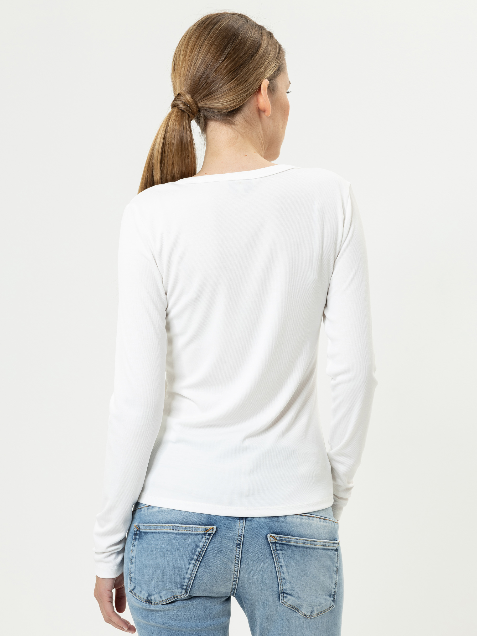 Eine Frau mit hellbraunem Haar in einem niedrigen Pferdeschwanz ist von hinten zu sehen. Sie trägt ein langärmeliges weißes Hemd und blaue Jeans mit sichtbaren Gesäßtaschen und steht vor einem schlichten weißen Hintergrund.