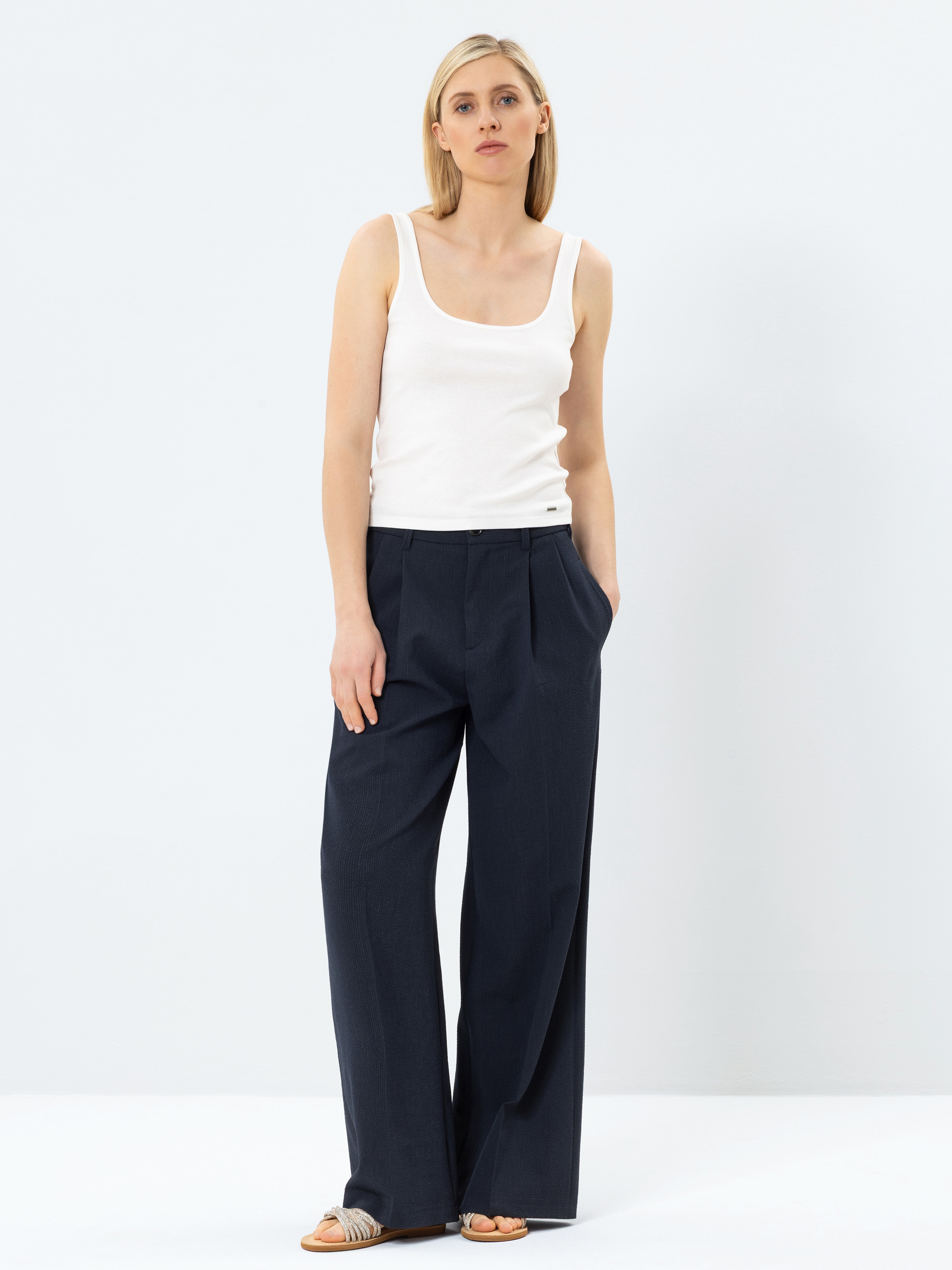 Eine Frau steht vor einem schlichten, hellen Hintergrund. Sie trägt ein weißes, ärmelloses Tanktop, eine dunkle, weit geschnittene Hose und Sandalen. Sie hat glattes blondes Haar und einen neutralen Gesichtsausdruck, eine Hand steckt in ihrer Tasche.