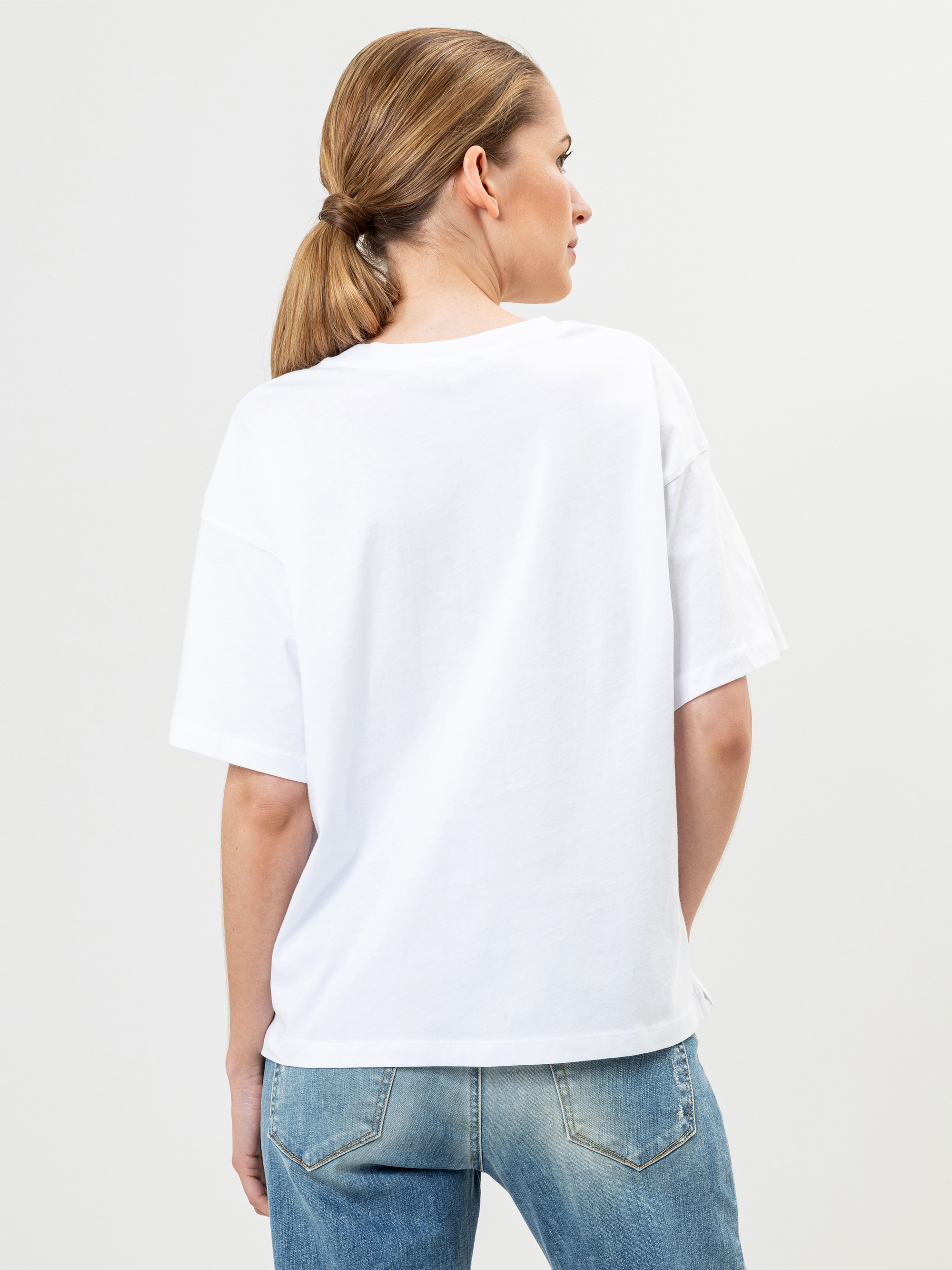 Eine Frau mit hellbraunem Haar in einem niedrigen Pferdeschwanz trägt ein lockeres weißes T-Shirt und hellblaue Jeans. Sie steht mit dem Rücken zur Kamera vor einem schlichten hellen Hintergrund.