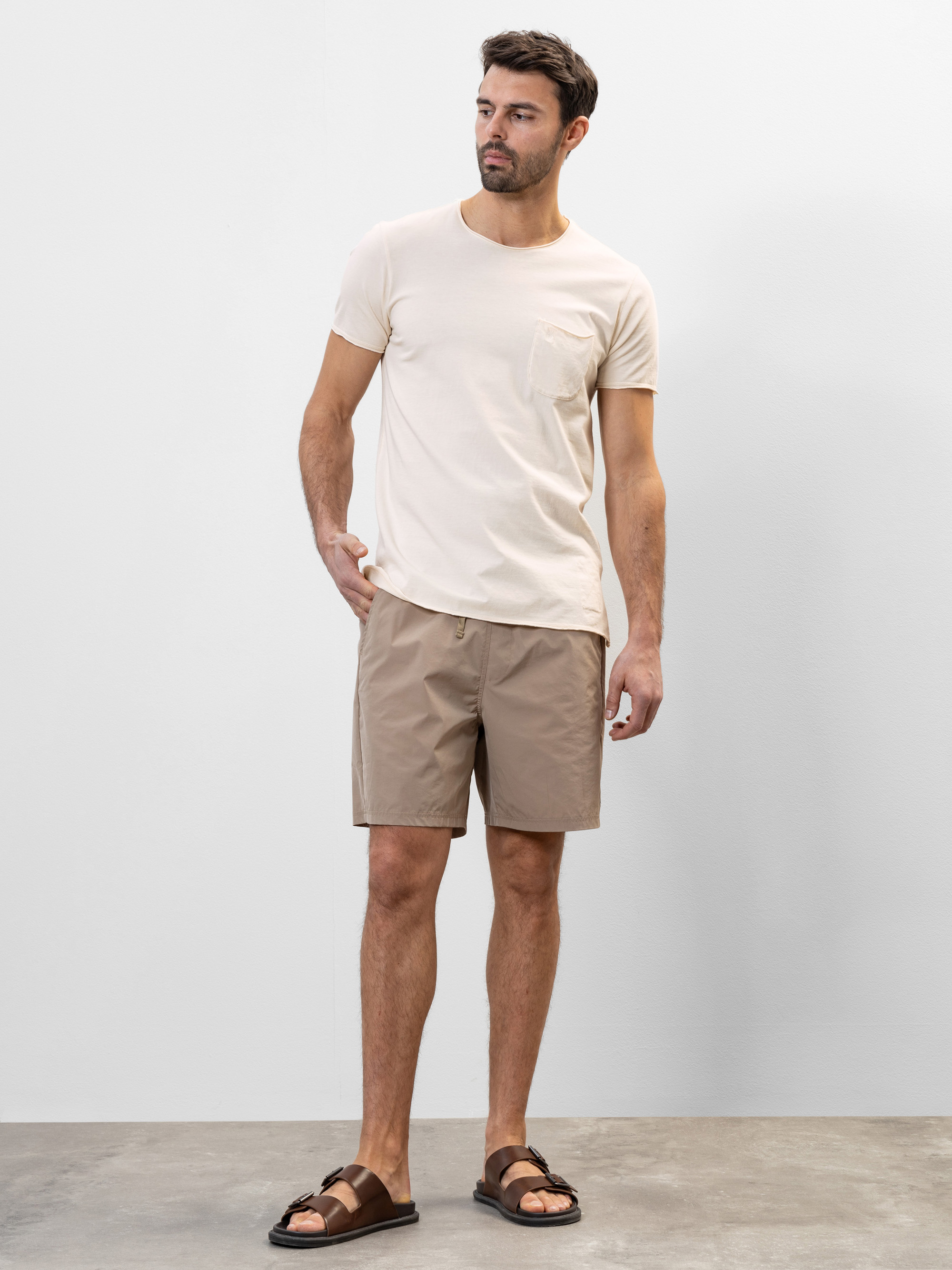 Ein Mann steht vor einer schlichten, hellgrauen Wand. Er trägt ein beiges T-Shirt mit einer Tasche, hellbraune Shorts und braune Sandalen. Er hat eine entspannte Körperhaltung und eine Hand in der Tasche.