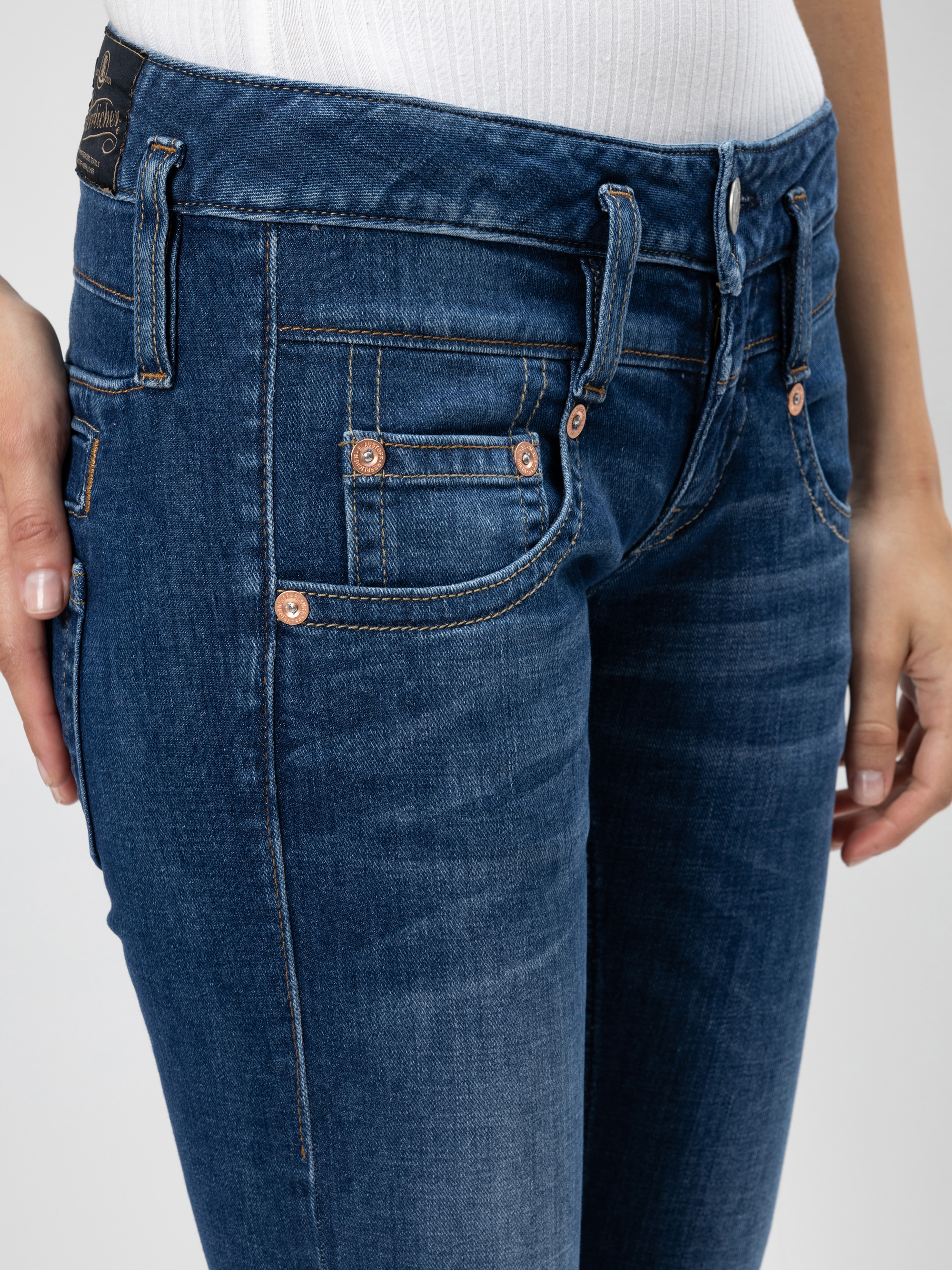 Nahaufnahme einer Person, die eine eng anliegende blaue Jeans und ein weißes geripptes Oberteil trägt, wobei eine Hand an der Seite ruht und der andere Arm teilweise sichtbar ist. Die Jeans hat kupferne Nieten und detaillierte Nähte.