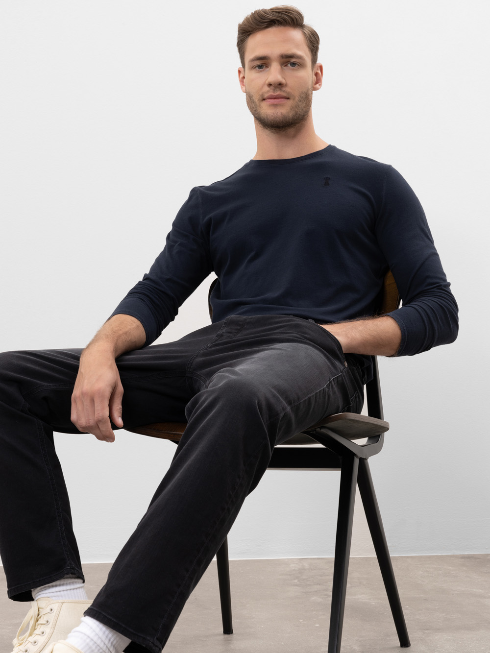 Ein Mann mit hellbraunem Haar, einem marineblauen langärmeligen Hemd, schwarzen Jeans und weißen Turnschuhen sitzt lässig auf einem Stuhl vor einem schlichten hellen Hintergrund und blickt selbstbewusst in die Kamera.