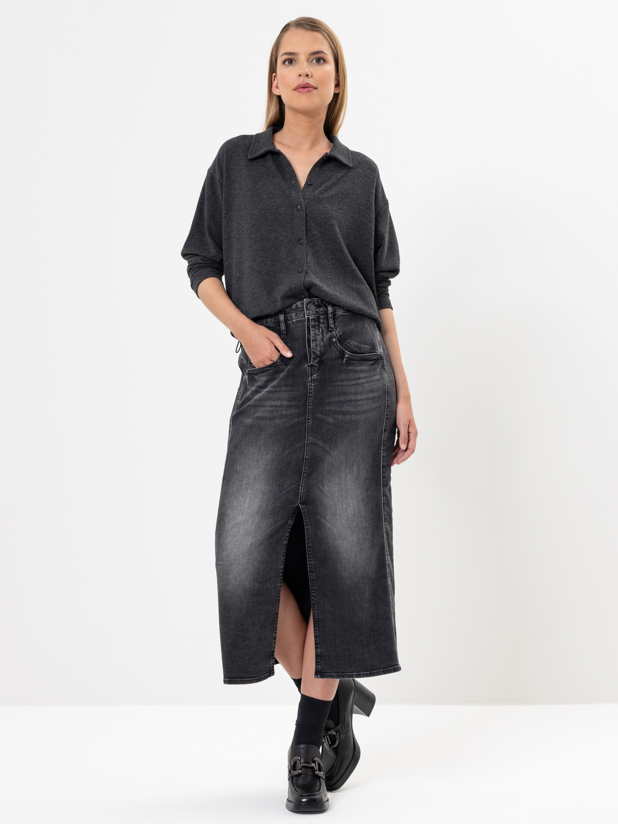 Eine Frau steht vor einem weißen Hintergrund und trägt ein dunkelgraues Button-up-Hemd und einen hochtaillierten, schwarzen Jeans-Midirock mit einem Schlitz an der Vorderseite, gepaart mit schwarzen Socken und schwarzen Schuhen. Sie hat eine Hand in ihr