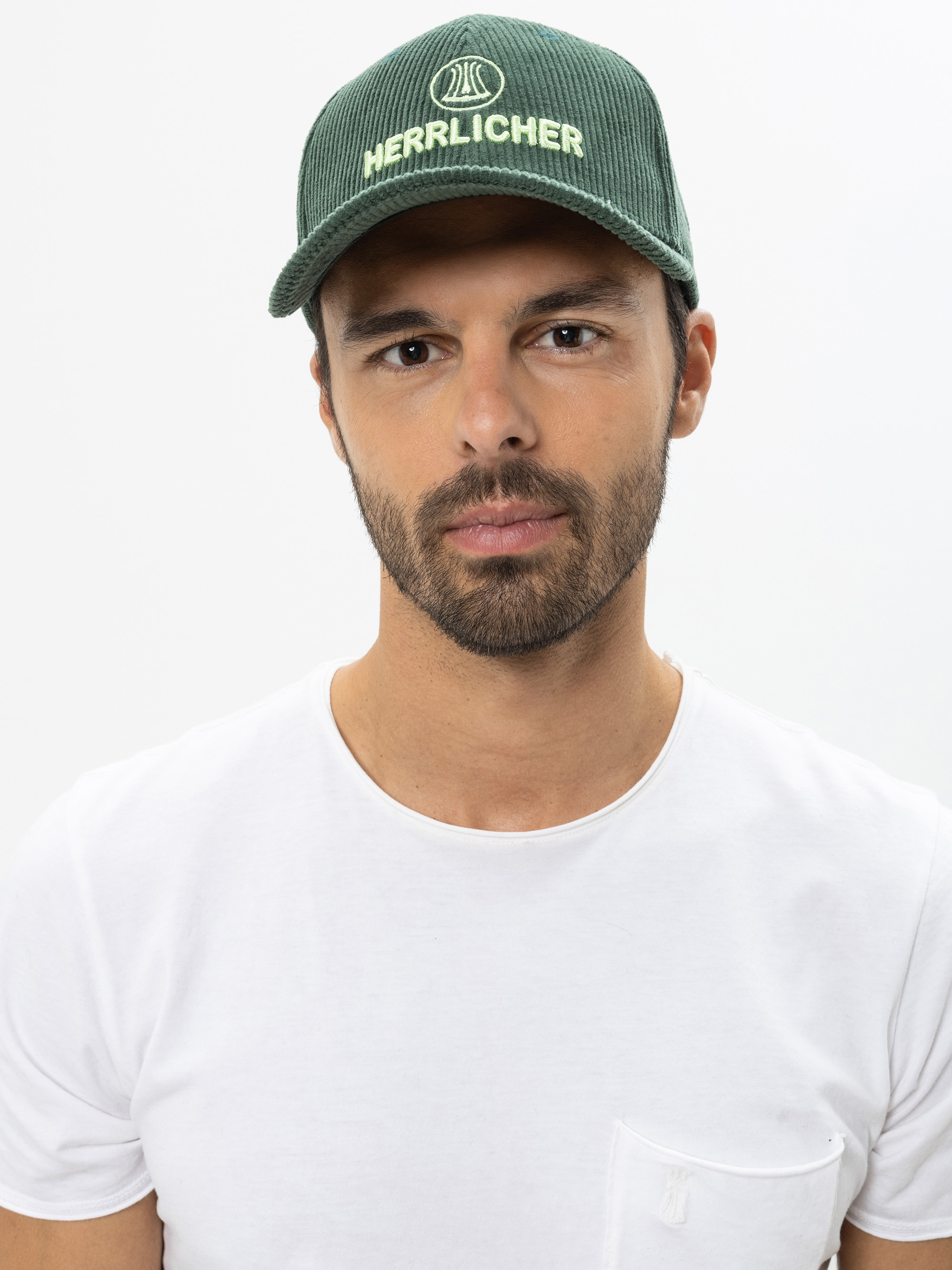 Ein Mann mit kurzem Bart und Schnurrbart, der eine grüne Kappe mit dem aufgestickten Wort Herrlicher und ein schlichtes weißes T-Shirt trägt, steht vor einem weißen Hintergrund.