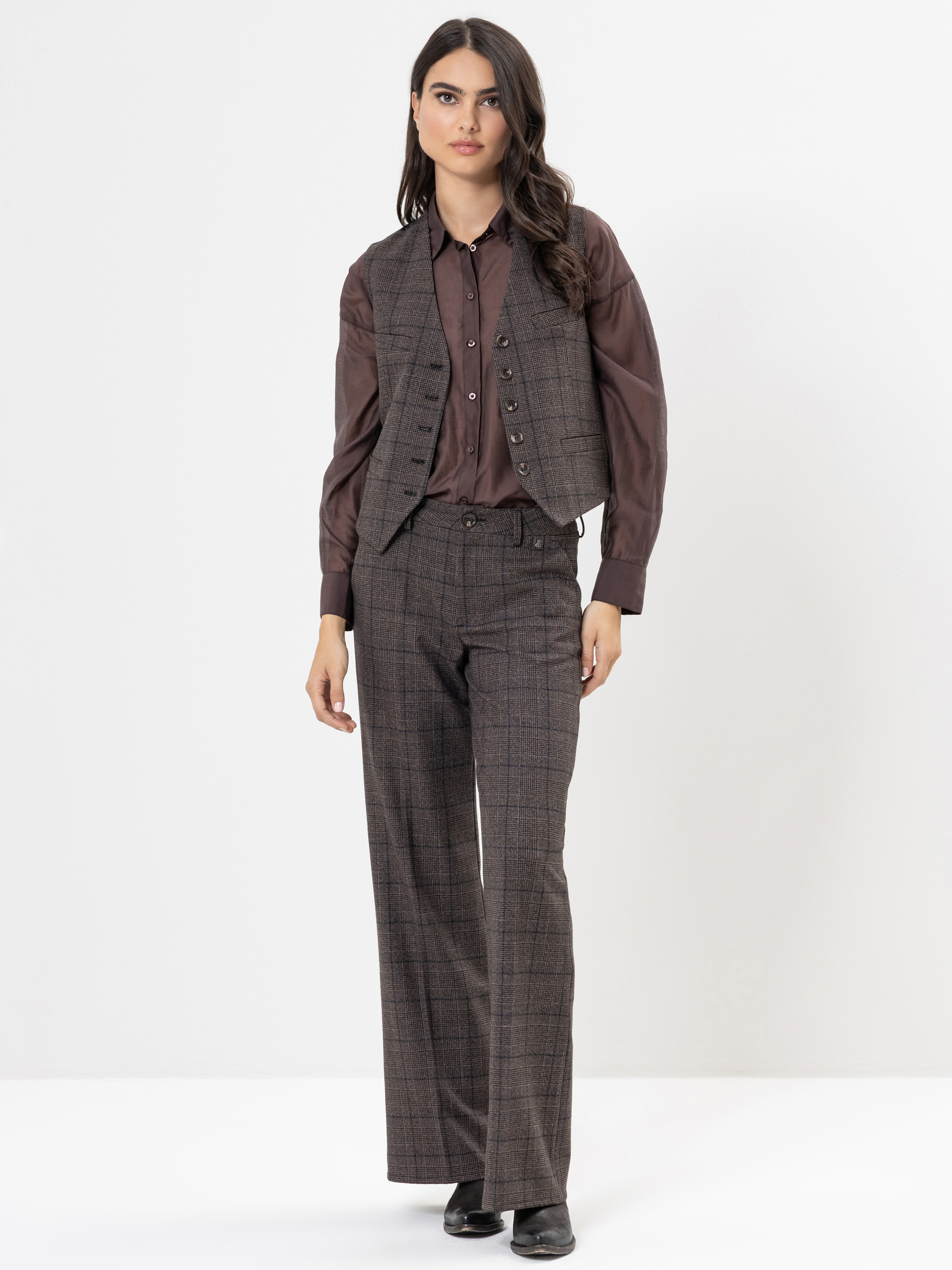 Eine Frau steht vor einem weißen Hintergrund und trägt eine braun karierte Weste und eine dazu passende Hose, gepaart mit einem schokoladenbraunen Button-up-Hemd und schwarzen Schuhen. Sie hat langes dunkles Haar und einen neutralen Gesichtsausdruck.