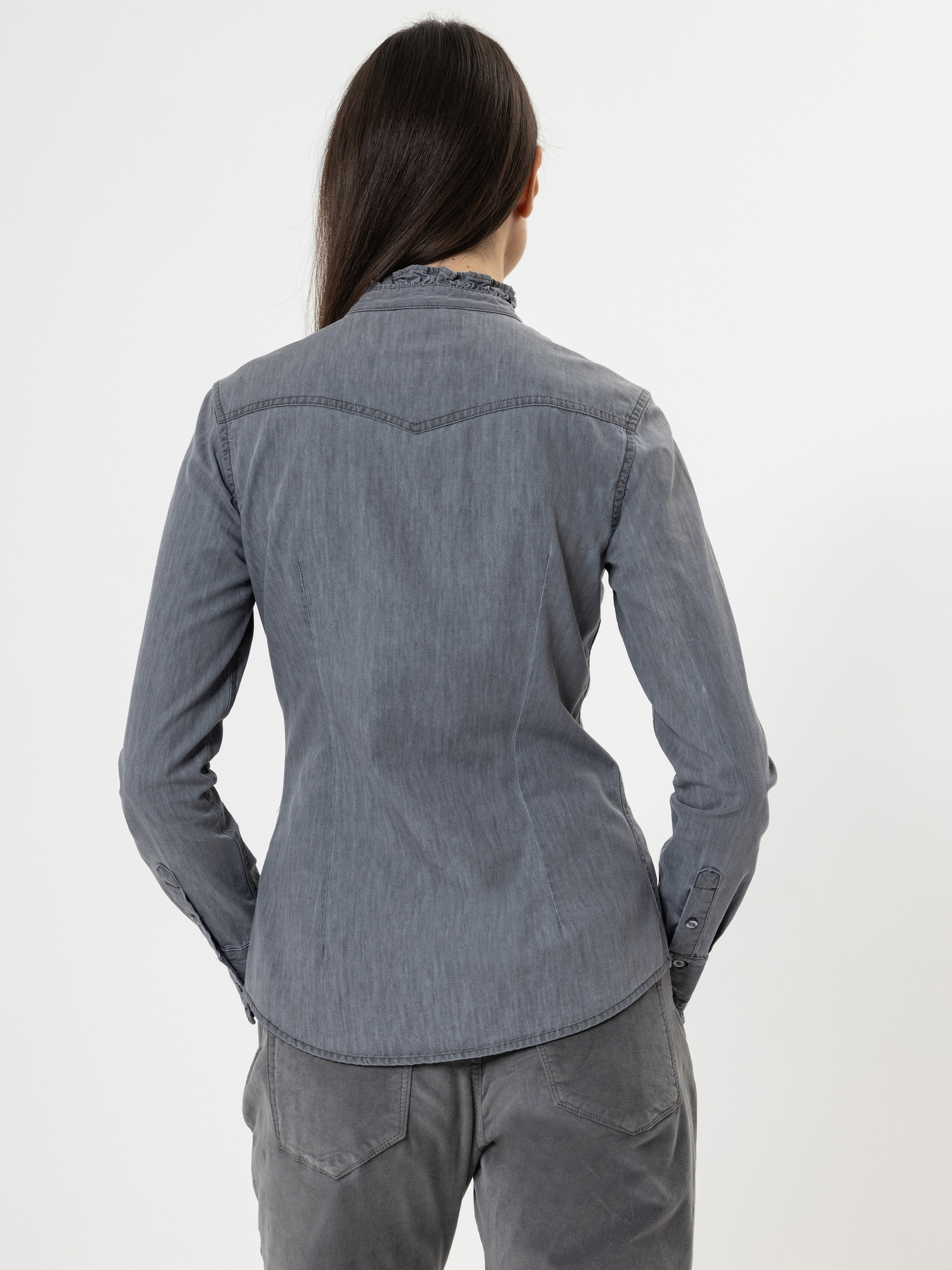 Eine Person mit langen dunklen Haaren, von hinten gesehen, trägt ein graues, geknöpftes Jeanshemd und eine passende graue Hose vor einem schlichten weißen Hintergrund.