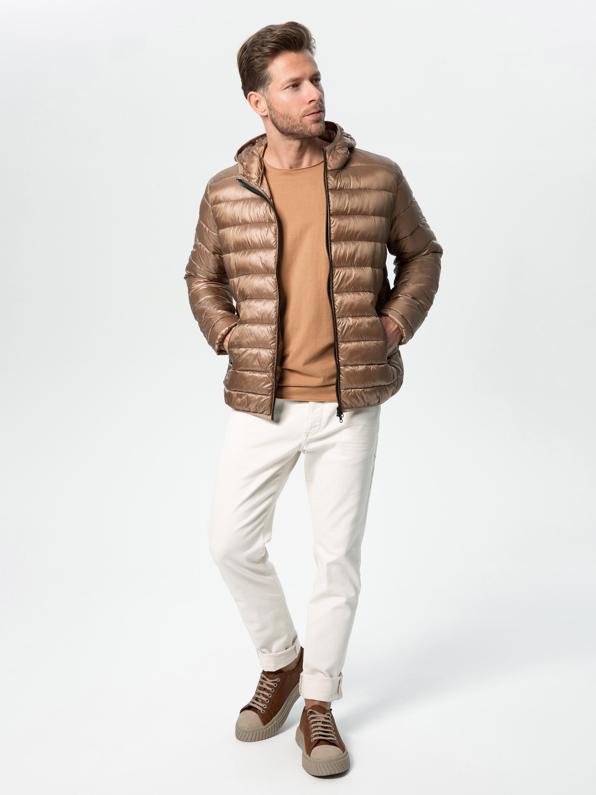 Ein Mann, der eine glänzende braune Pufferjacke, einen hellbraunen Pullover, eine weiße Hose und braune Turnschuhe trägt, steht mit den Händen in den Taschen und blickt vor einem schlichten hellen Hintergrund zur Seite.