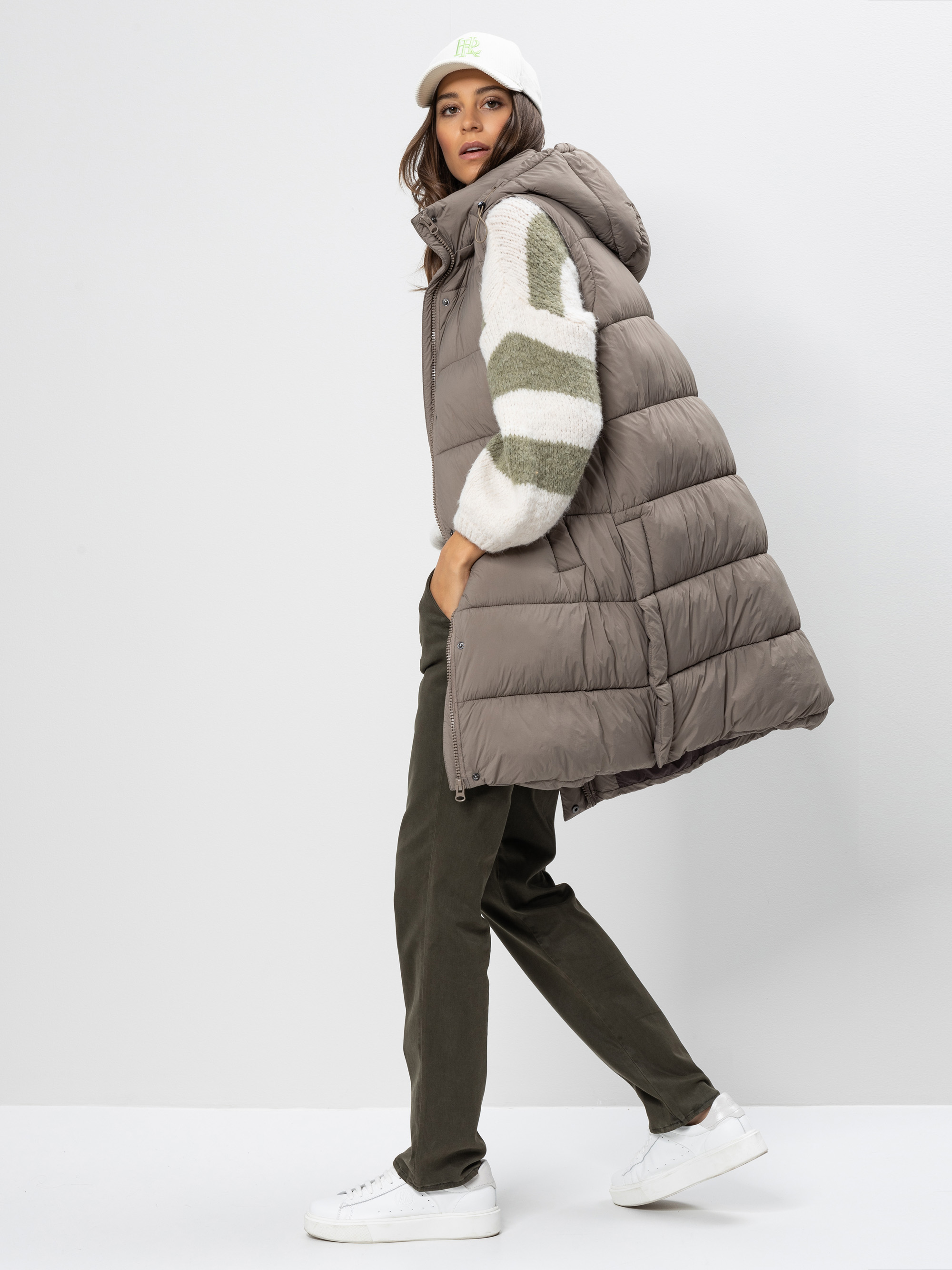 Eine Frau in einer langen, taupefarbenen Pufferweste, einem gestreiften Pullover, einer dunklen Hose und weißen Turnschuhen steht seitwärts und wendet ihren Kopf der Kamera zu. Sie trägt eine weiße Baseballmütze und posiert vor einem schlichten weiß