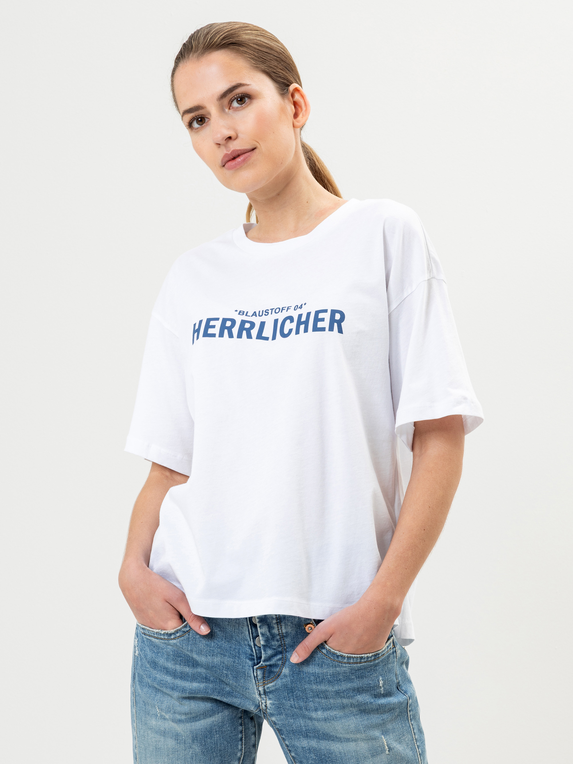 Eine Frau mit hellbraunem Haar in einem niedrigen Pferdeschwanz trägt ein lockeres weißes T-Shirt mit dem Aufdruck HERRLICHER in Blau und hellblaue Jeans. Sie steht mit den Händen in den Taschen vor einem einfarbigen Hintergrund.