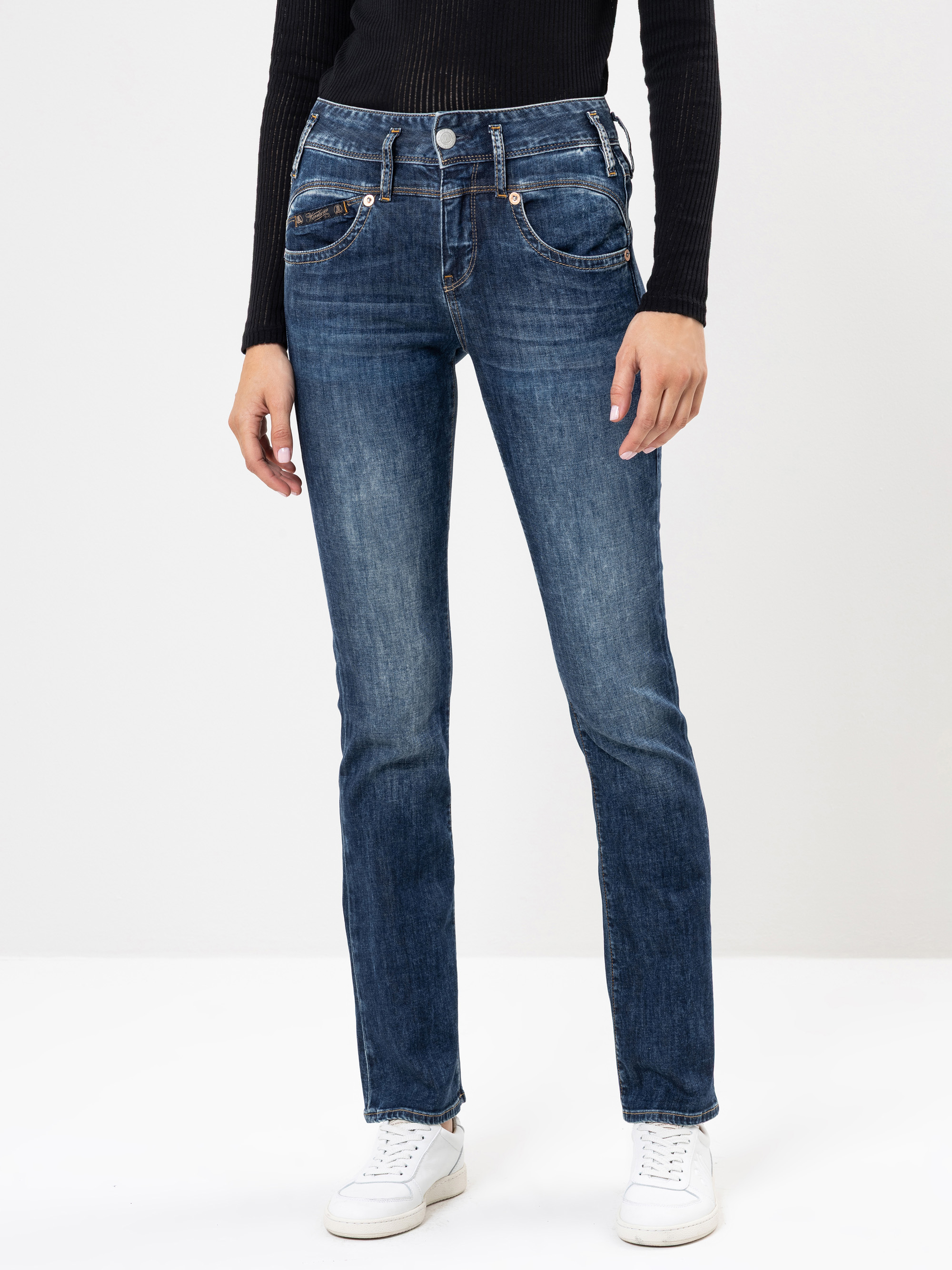 Eine Person, die ein schwarzes geripptes langärmeliges Oberteil, eine blaue Jeans mit geraden Beinen und weiße Turnschuhe trägt, steht vor einem schlichten weißen Hintergrund.