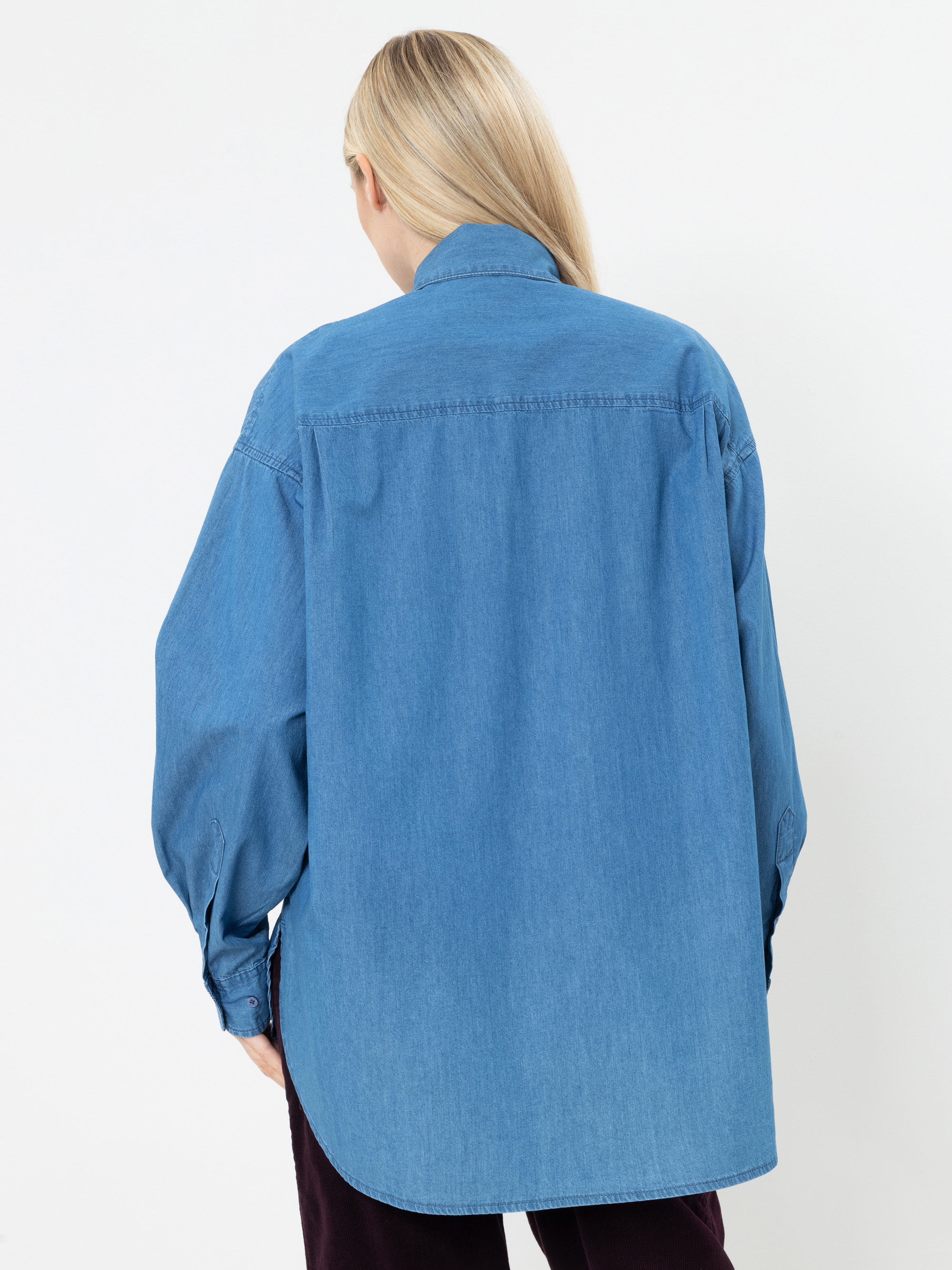 Eine Person mit langen blonden Haaren, die ein übergroßes blaues Jeanshemd trägt, steht mit dem Rücken zur Kamera vor einem schlichten weißen Hintergrund.