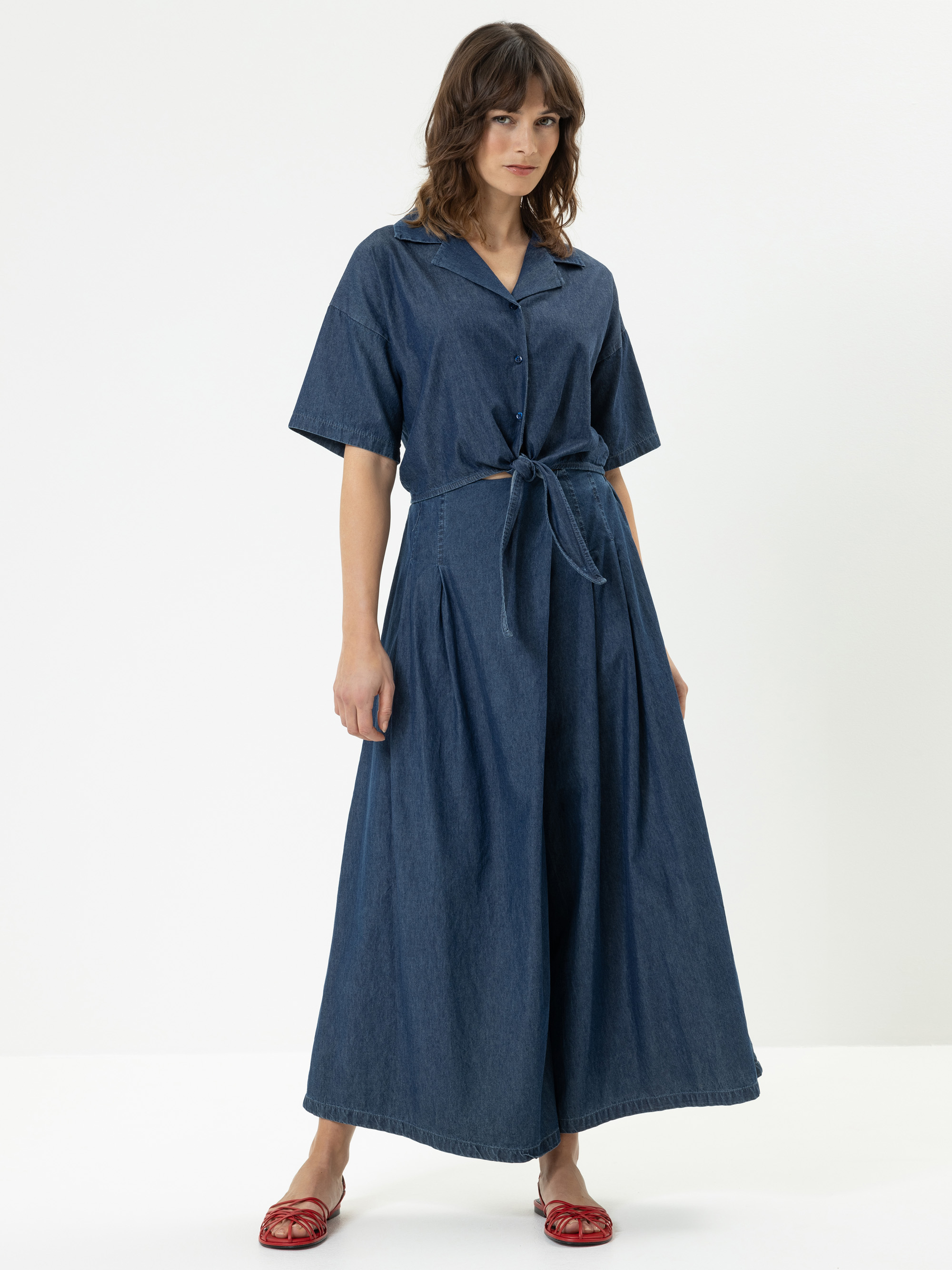 Eine Frau steht vor einem einfarbig weißen Hintergrund und trägt einen Jeansoverall mit weitem Bein, kurzen Ärmeln, einem Kragen und einem Bindeband in der Taille. Sie trägt außerdem rote flache Schuhe.