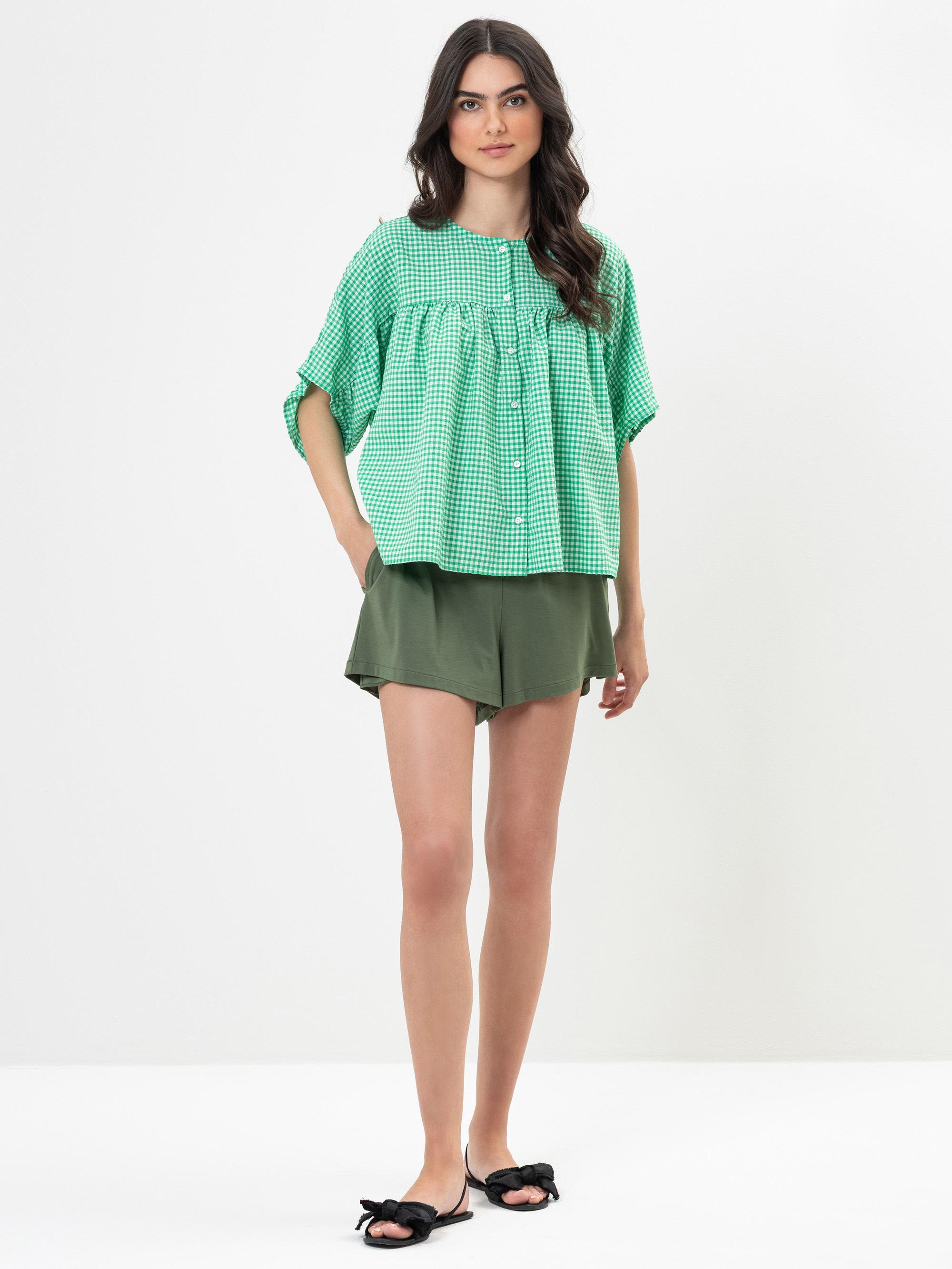 Frau, die vor einem schlichten weißen Hintergrund steht und ein lockeres grünes, kariertes Oberteil, olivgrüne Shorts und schwarze Slide-Sandalen mit Schleifen trägt. Sie hat langes, gewelltes dunkles Haar und einen neutralen Gesichtsausdruck.