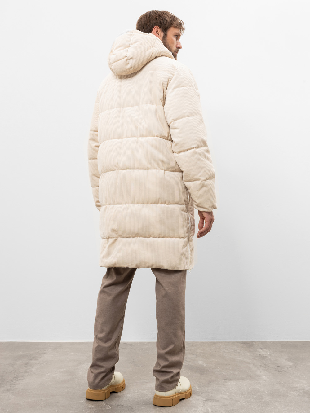 Ein Mann steht mit dem Gesicht zur Wand, in einer langen, cremefarbenen Pufferjacke mit Kapuze, einer taupefarbenen Hose und hellbeigen Schuhen, vor einer schlichten weißen Wand und einem grauen Boden.
