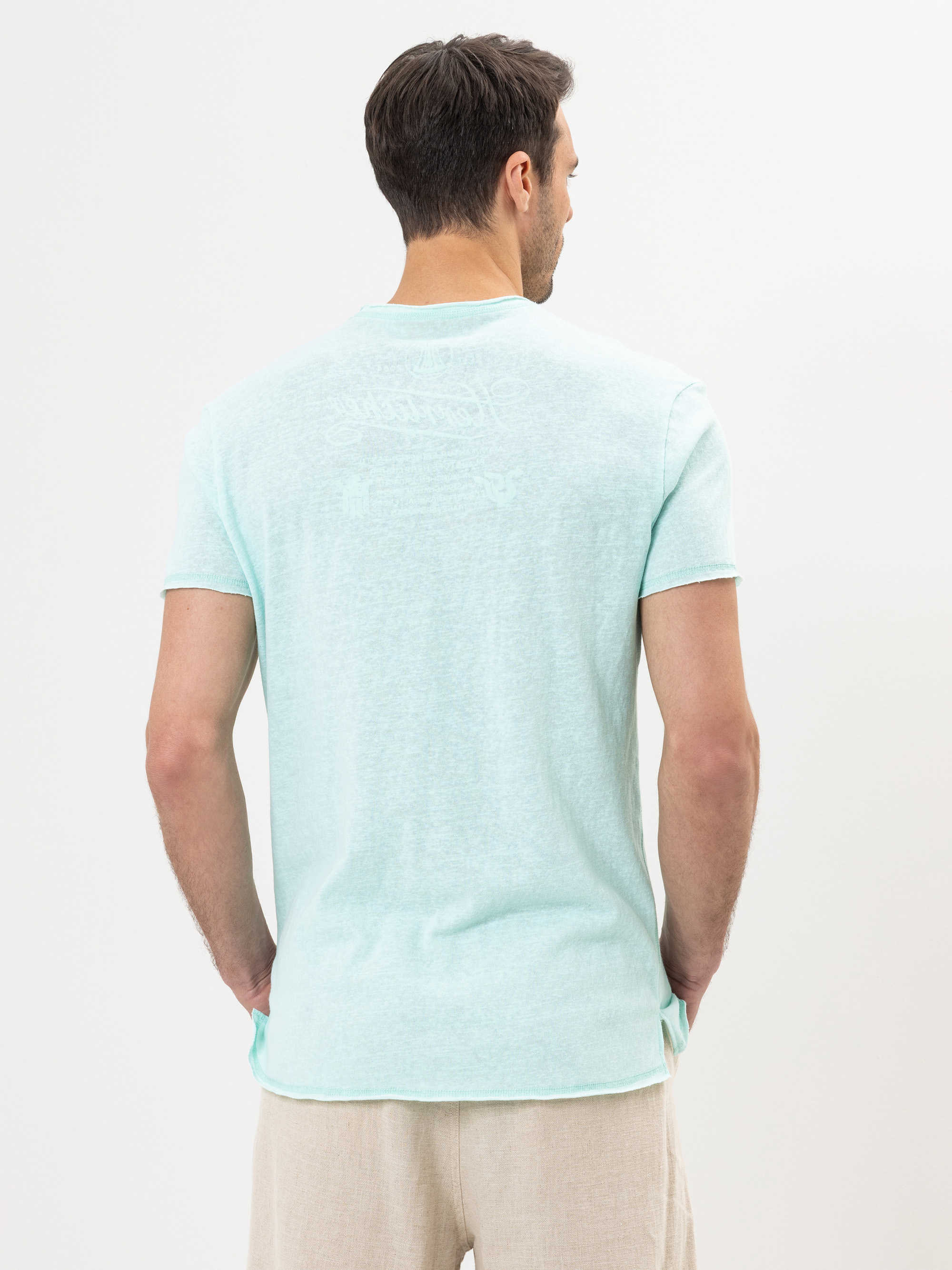 Ein Mann mit kurzen dunklen Haaren ist von hinten zu sehen. Er trägt ein hellblaues kurzärmeliges T-Shirt und beigefarbene Shorts und steht vor einem schlichten weißen Hintergrund.