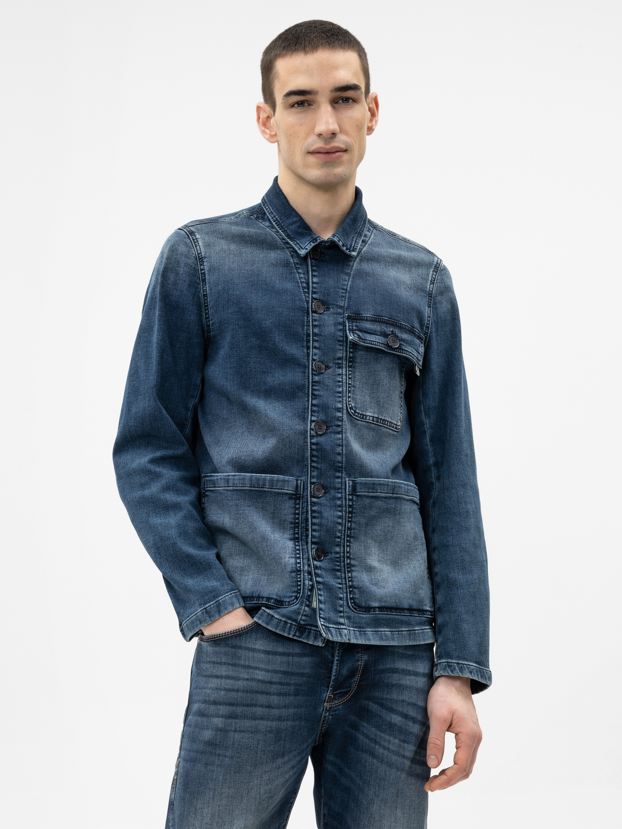 Eine Person, die eine blaue Jeansjacke mit großen Vordertaschen und dazu passende blaue Jeans trägt, steht vor einem schlichten weißen Hintergrund. Sie hat kurzes dunkles Haar und einen neutralen Gesichtsausdruck.