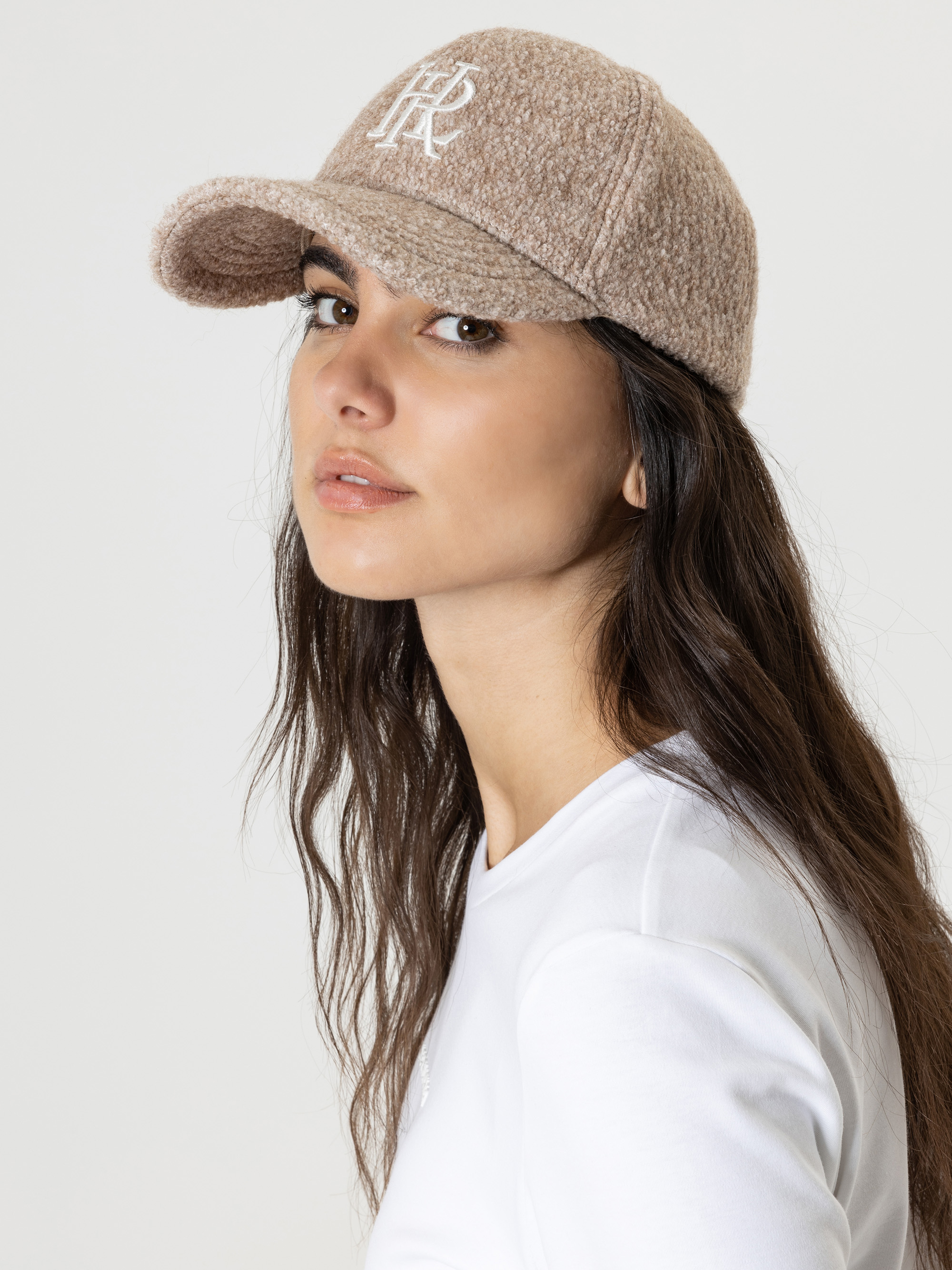 Eine Frau mit langen braunen Haaren, die eine beigefarbene Baseballkappe mit weißen aufgestickten Buchstaben und ein weißes Hemd trägt, posiert vor einem schlichten hellen Hintergrund.