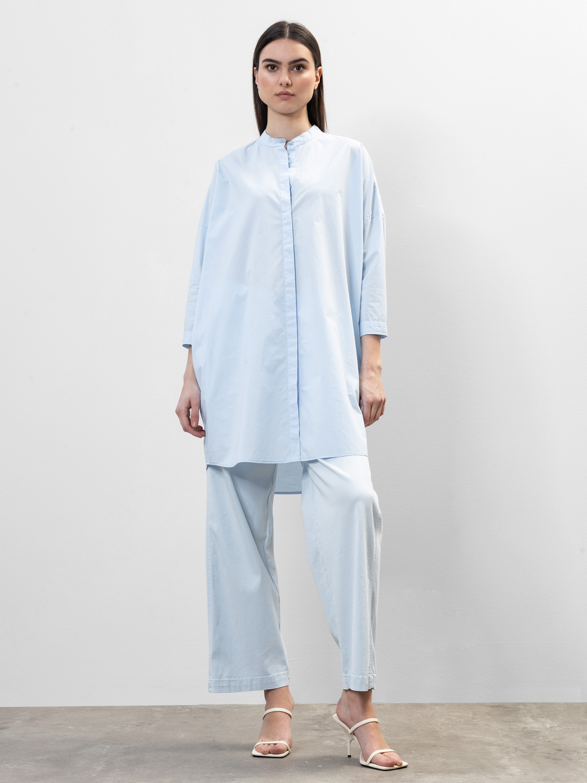 Eine Frau steht vor einer schlichten weißen Wand und trägt eine locker sitzende, hellblaue Tunika mit weiten Ärmeln und eine dazu passende Hose mit weiten Beinen. Sie hat glattes dunkles Haar und trägt weiße, hochhackige Sandalen.