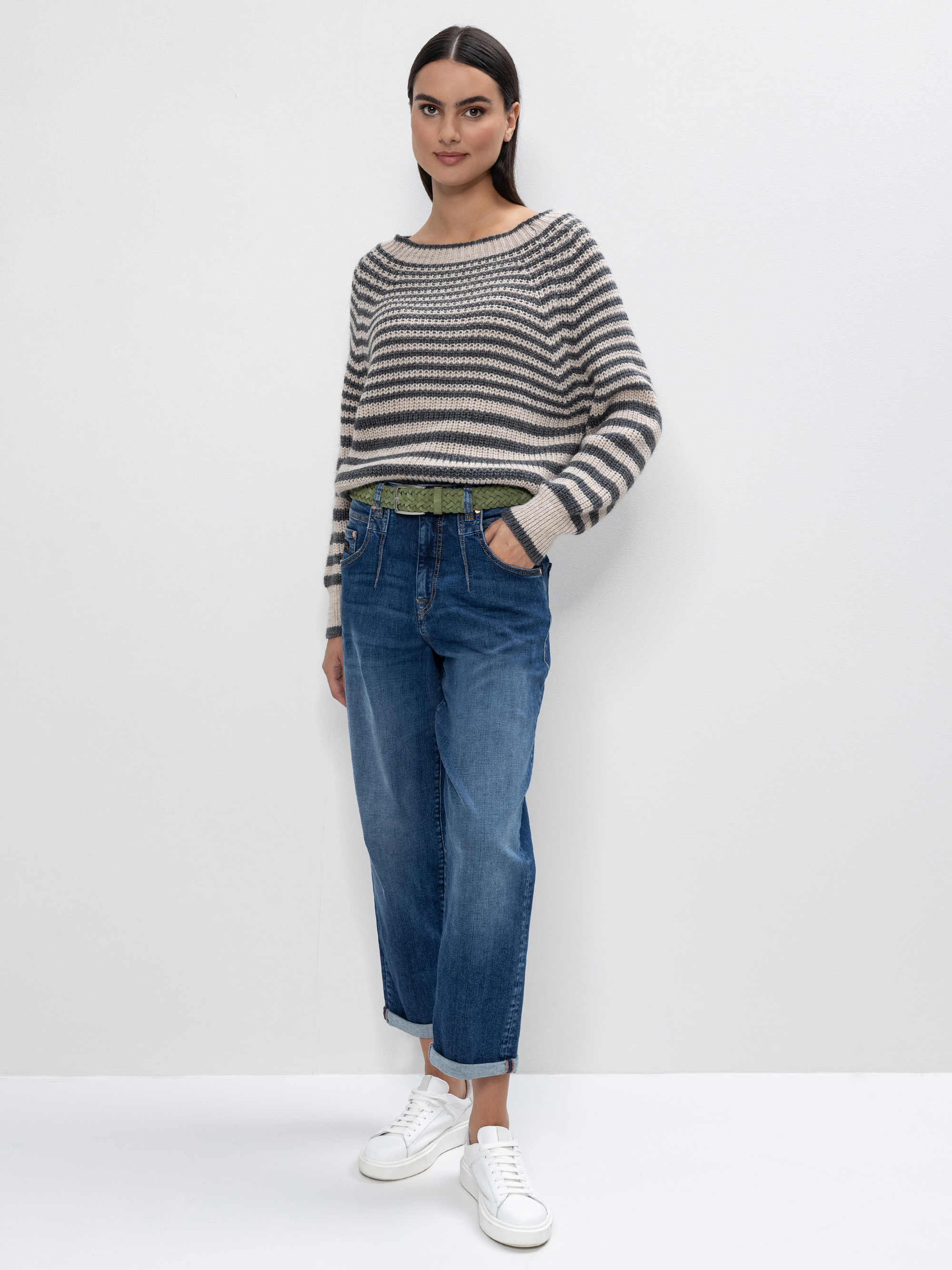 Eine Frau steht vor einer schlichten weißen Wand und trägt einen gestreiften langärmeligen Pullover, blaue Jeans und weiße Turnschuhe. Sie hat ihre linke Hand in der Hosentasche und schaut mit einem leichten Lächeln in die Kamera.