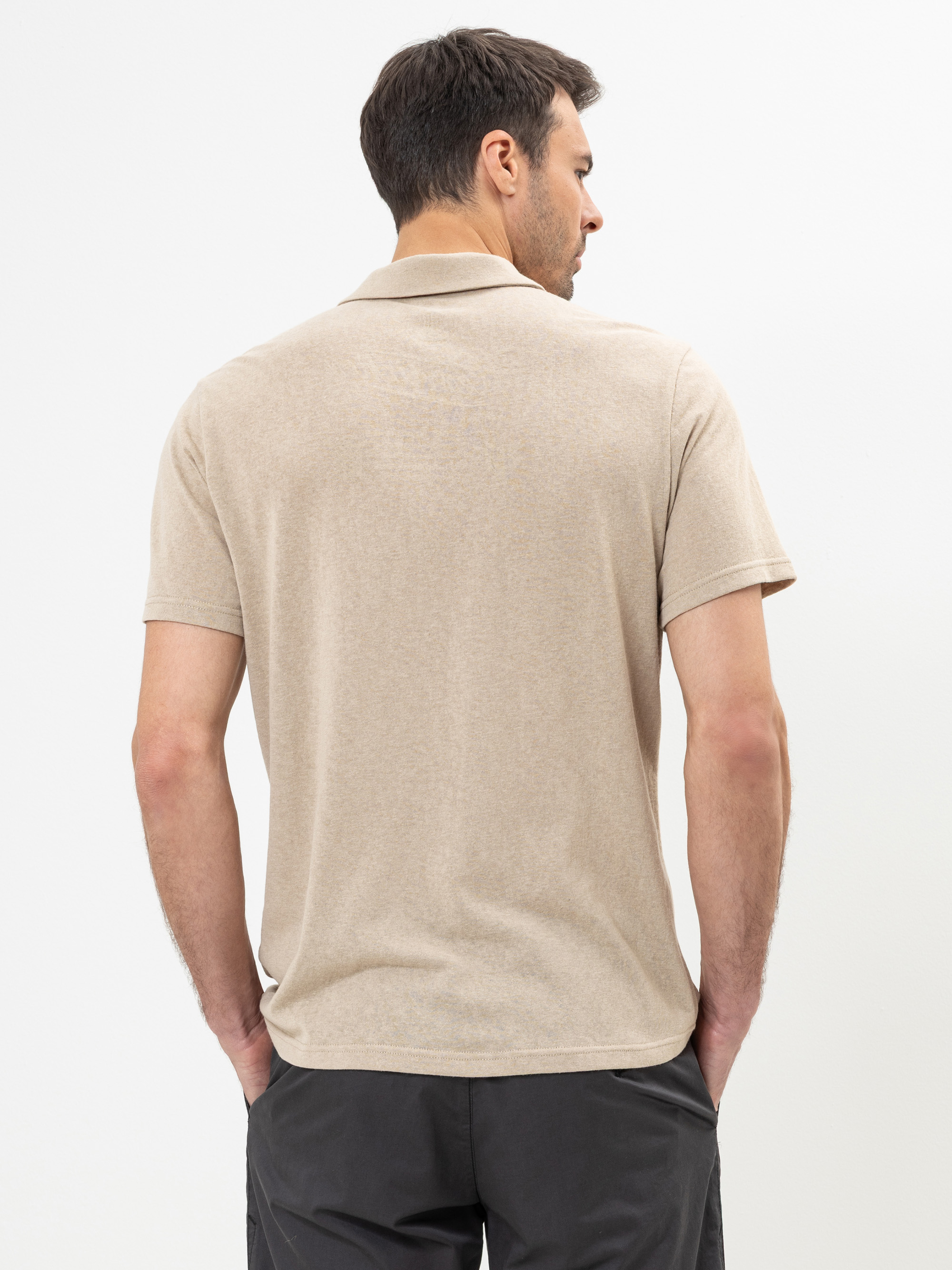 Ein Mann mit kurzen braunen Haaren steht mit dem Rücken zur Kamera, trägt ein beiges kurzärmeliges Polohemd und eine dunkle Hose vor einem schlichten weißen Hintergrund.