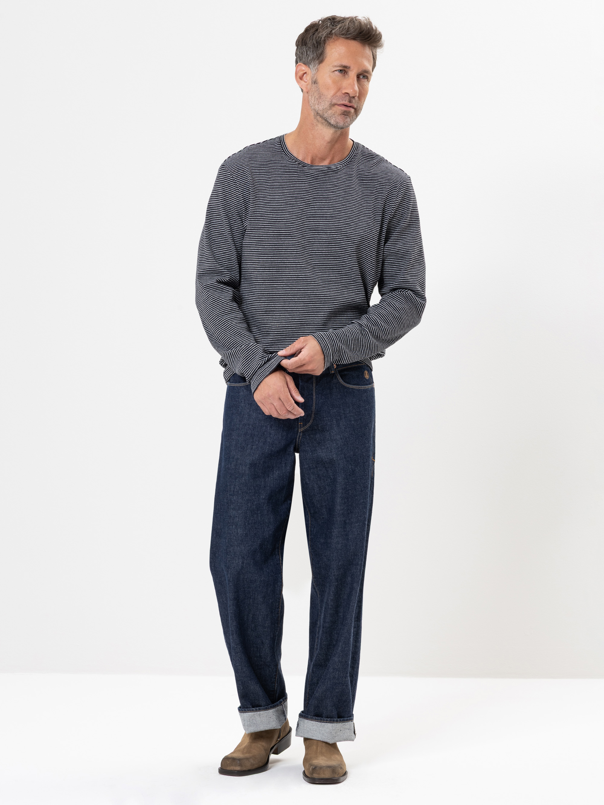 Ein Mann mit kurzen Haaren und Bartstoppeln trägt einen grauen strukturierten Pullover, eine locker sitzende dunkelblaue Jeans mit aufgerollten Bündchen und braune Schuhe und steht vor einem schlichten weißen Hintergrund.