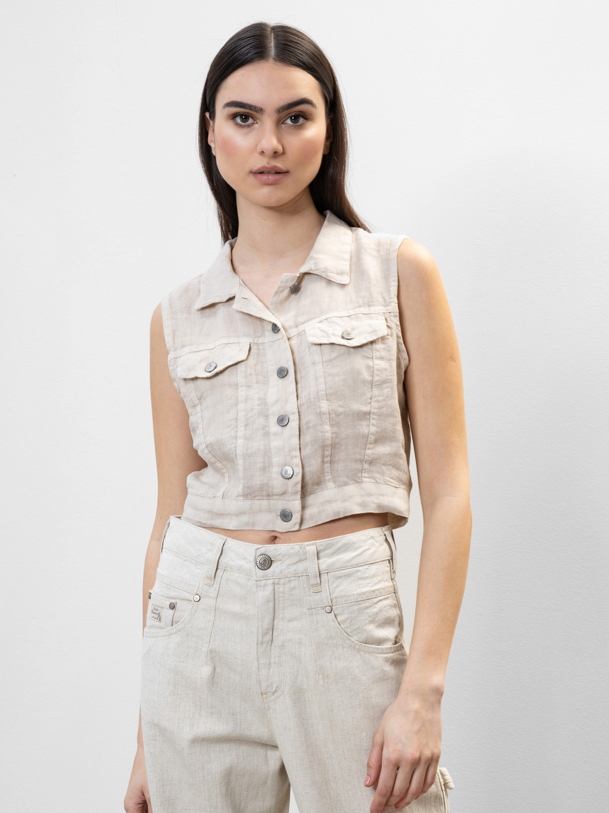 Eine Frau mit langen dunklen Haaren trägt ein ärmelloses, hellbeiges, geknöpftes Oberteil und eine dazu passende Hose mit hoher Taille vor einem schlichten hellen Hintergrund.