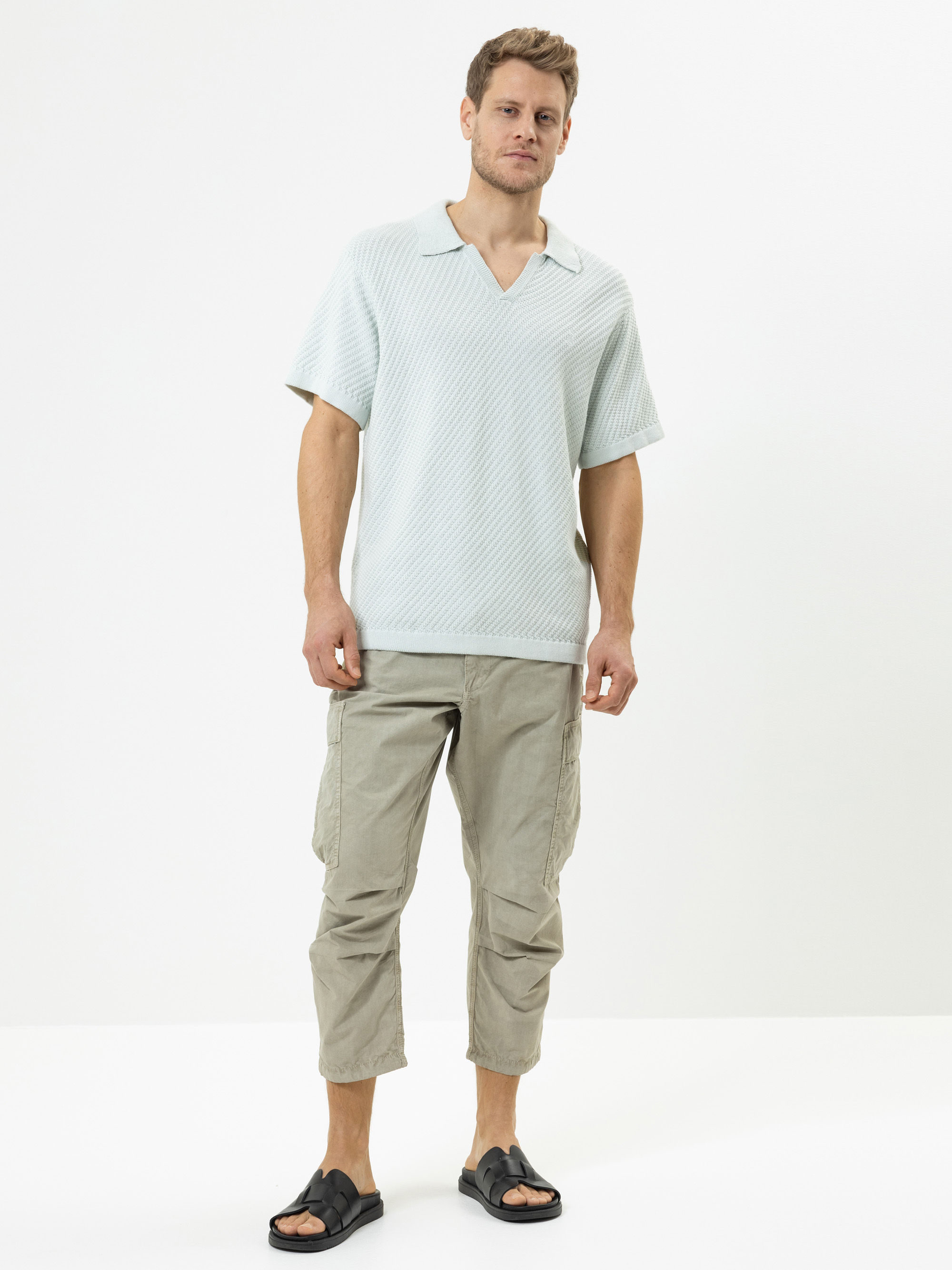 Ein Mann steht vor einem weißen Hintergrund und trägt ein hellgraues, kurzärmeliges Poloshirt, eine helle, khakifarbene Cargohose und schwarze Slide-Sandalen. Er blickt mit neutraler Miene nach vorne.