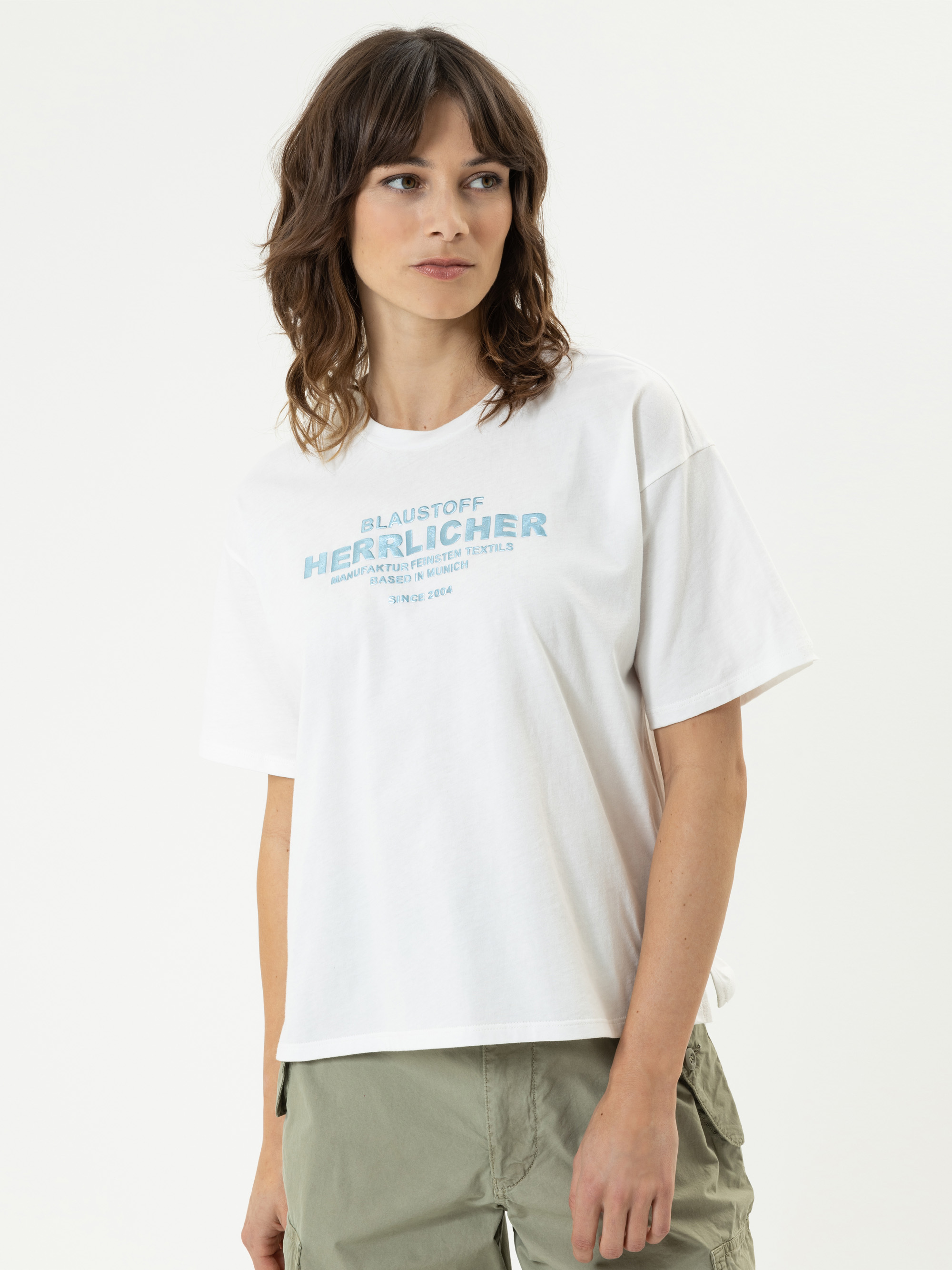 Eine Frau mit gewelltem braunem Haar, die ein weißes grafisches T-Shirt und eine hellgrüne Hose trägt, steht leicht zur Seite gewandt vor einem schlichten hellen Hintergrund.