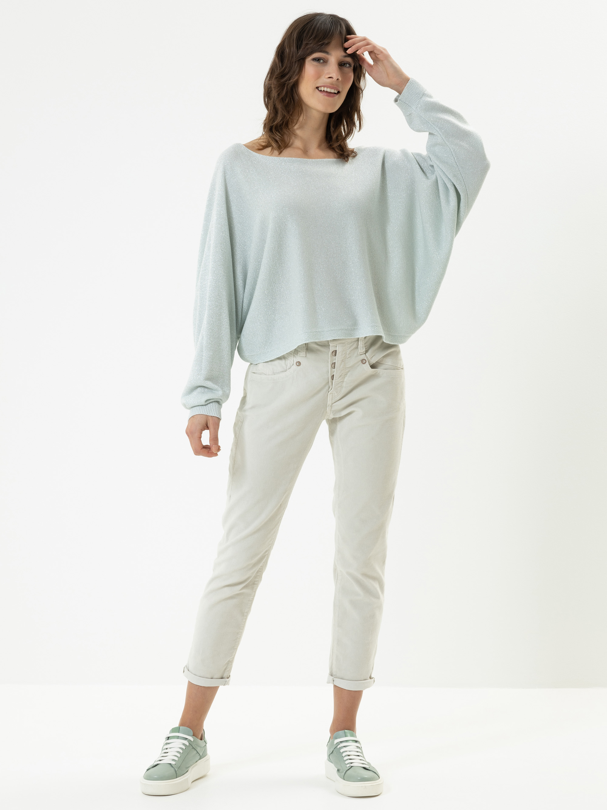 Eine Frau steht lächelnd in einem lockeren hellblauen Pullover, einer hellen Hose mit Bündchen und hellgrünen Turnschuhen vor einem schlichten weißen Hintergrund.