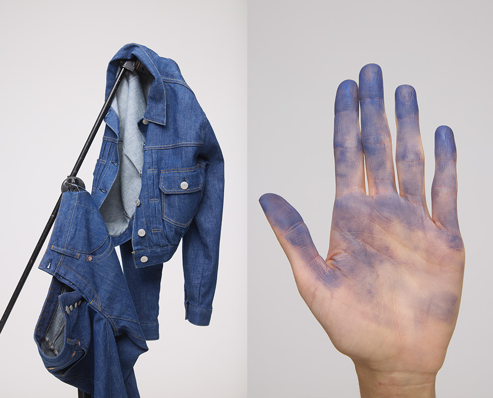 Eine Jeansjacke und eine Jeans hängen auf einem schwarzen Ständer vor einem weißen Hintergrund; daneben ist eine geöffnete Hand mit blauen Farbflecken auf Fingern und Handfläche zu sehen.