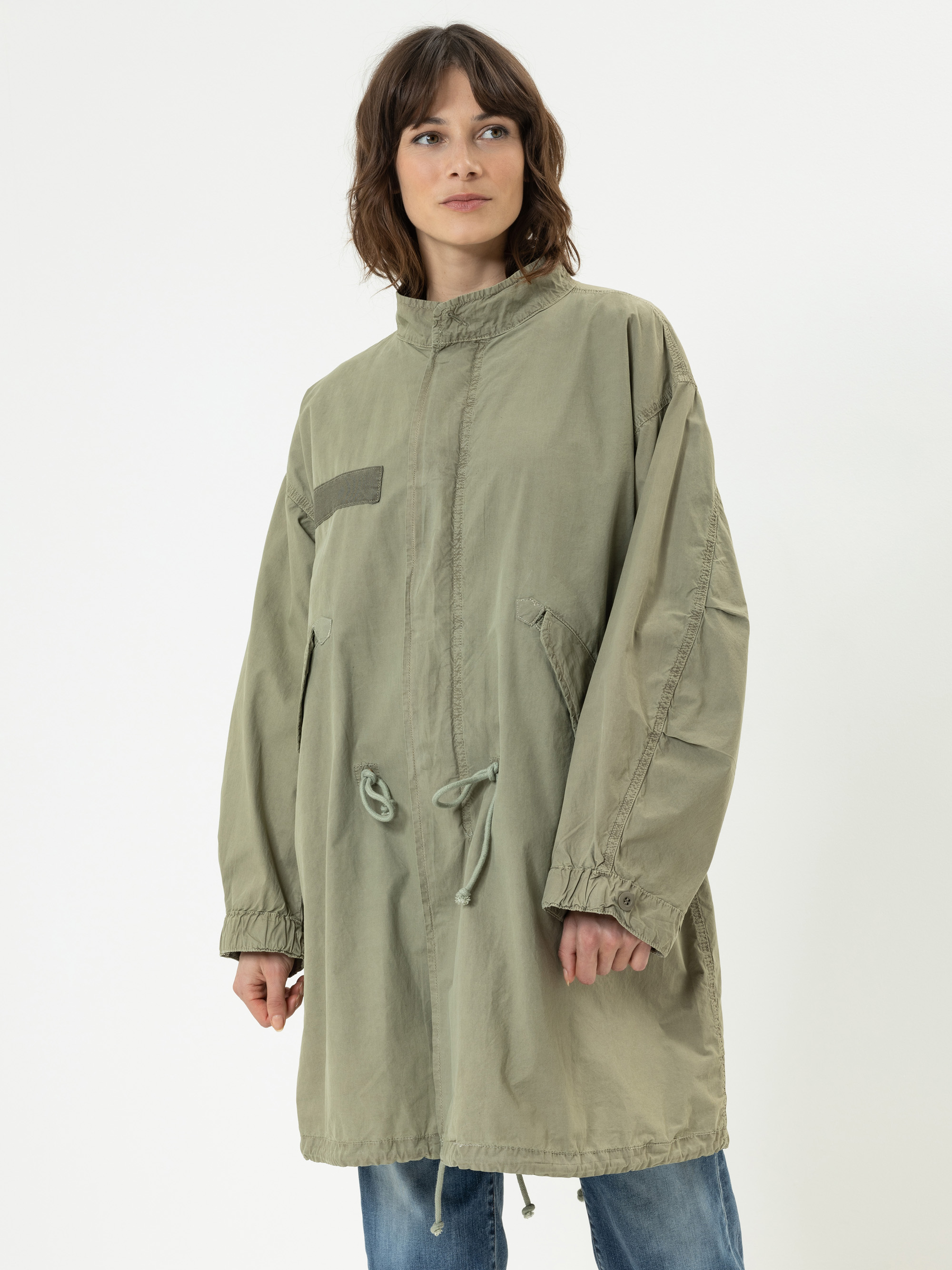 Eine Frau trägt eine locker sitzende, hellolivgrüne Parka-Jacke mit Kordelzug in der Taille, langen Ärmeln, hohem Kragen und zwei großen Vordertaschen. Sie steht vor einem schlichten weißen Hintergrund.