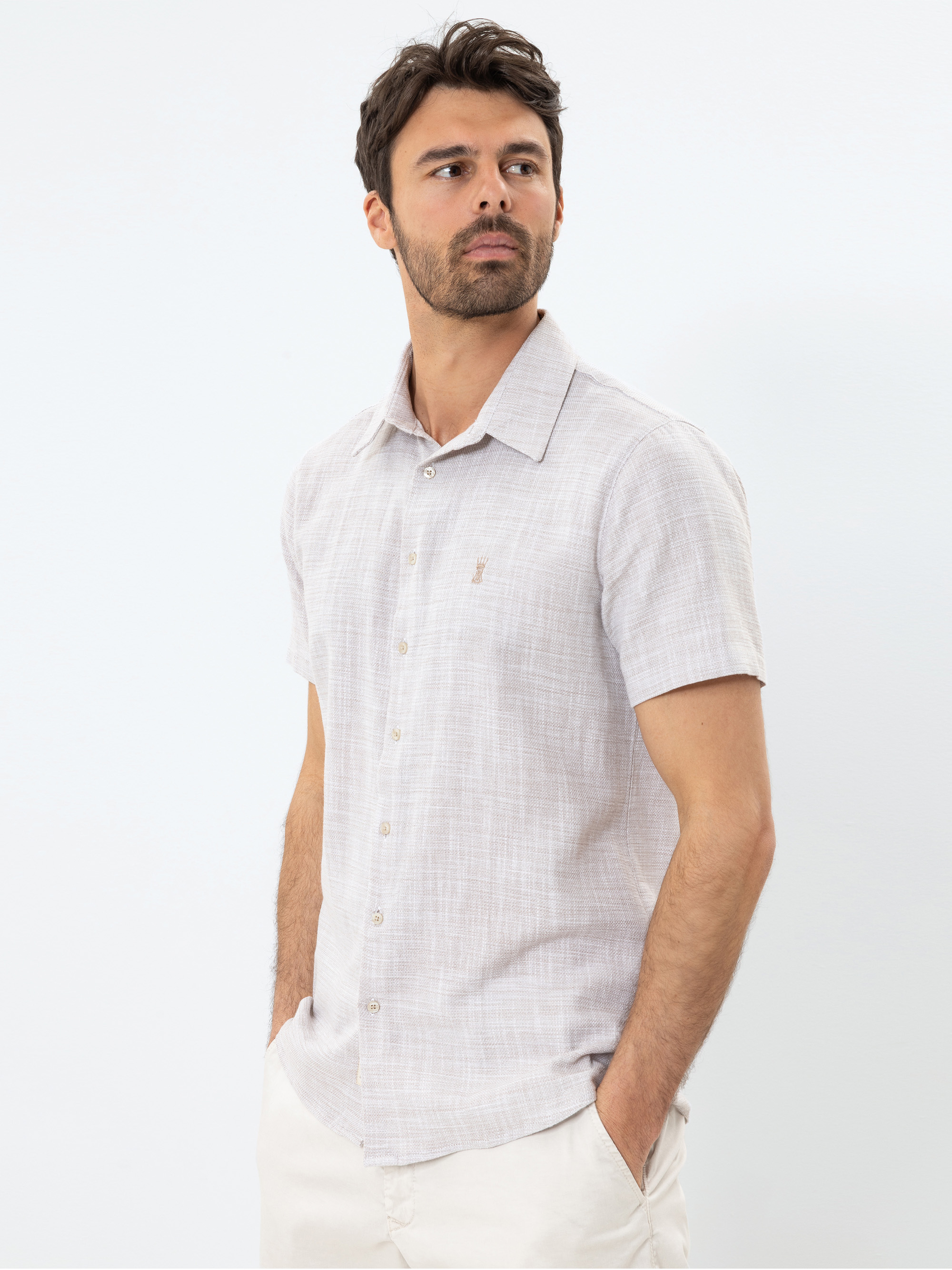 Ein Mann mit dunklem Haar und Bart trägt ein helles, kurzärmeliges Button-up-Hemd und eine helle Hose. Er steht mit den Händen in den Taschen und blickt zur Seite vor einem schlichten weißen Hintergrund.