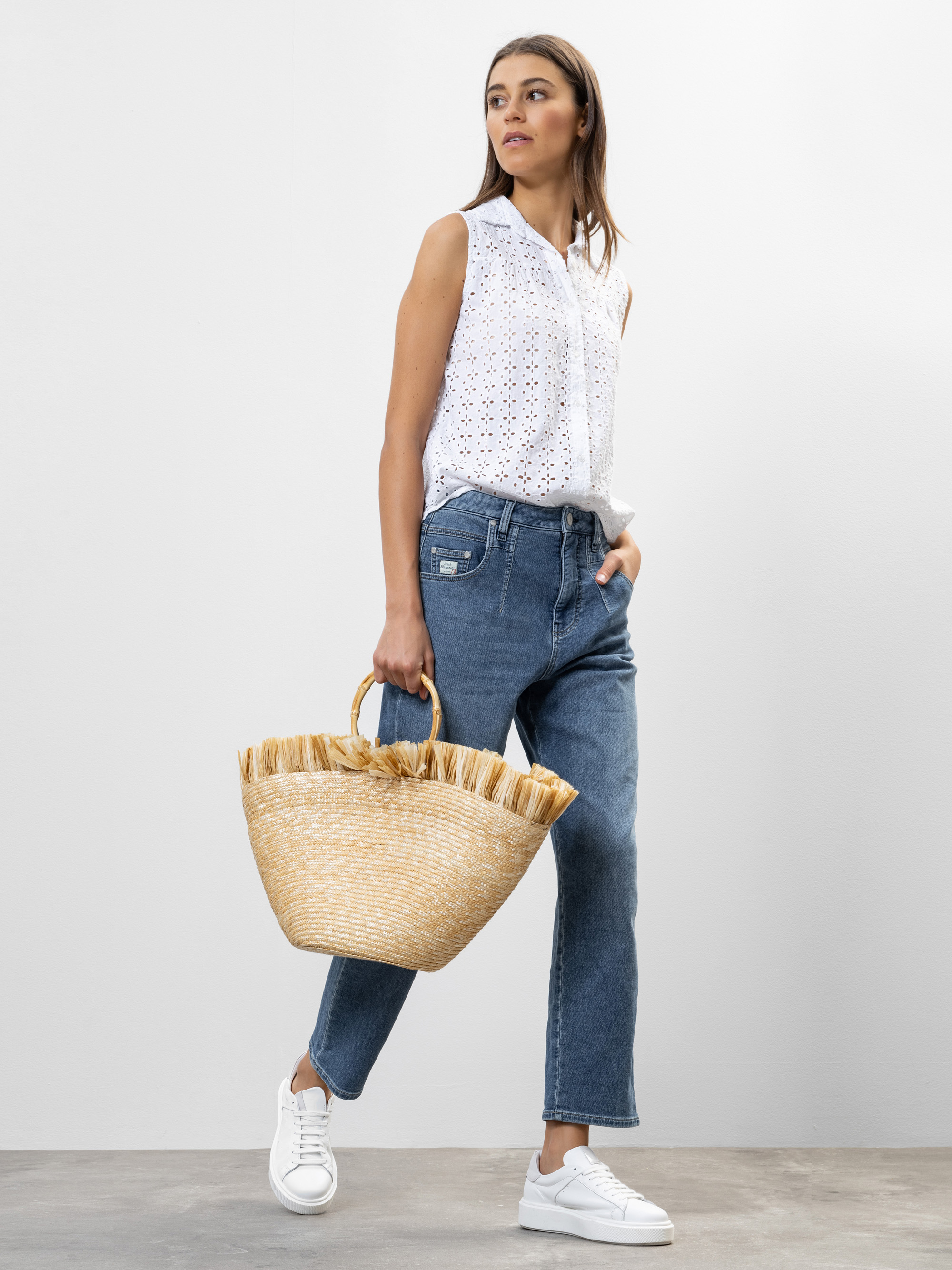 Eine Frau steht vor einem schlichten Hintergrund und trägt eine ärmellose weiße Bluse mit Ösen, blaue Jeans und weiße Turnschuhe. Sie hält eine Strohtasche mit Fransen in der Hand und hat eine Hand in der Tasche, wobei sie leicht zur Seite schaut.