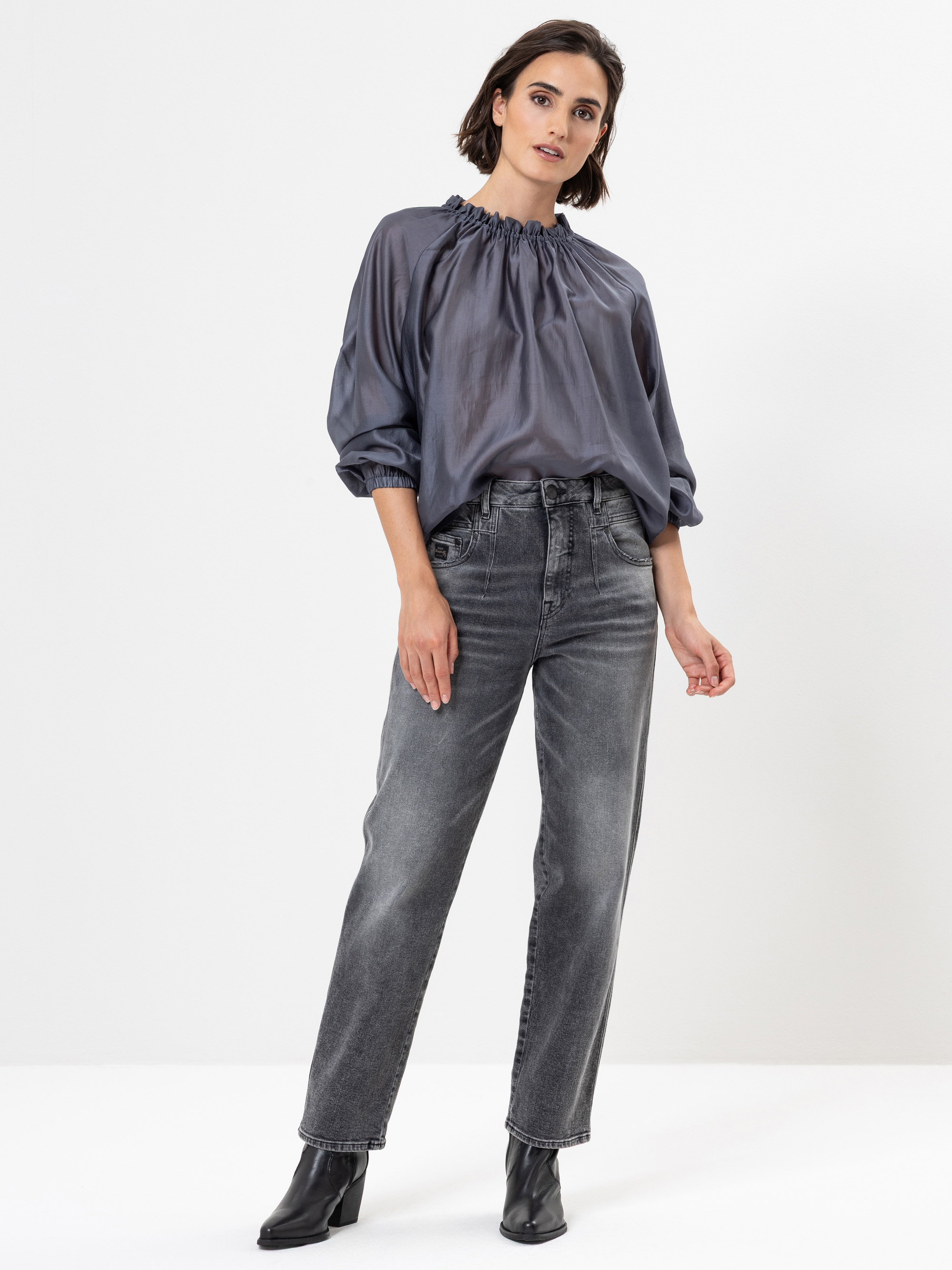 Eine Frau steht in einem Atelier und trägt eine glänzende graue Bluse mit gerafftem Ausschnitt, hochtaillierte, verwaschene schwarze Jeans und schwarze Ankle Boots. Sie hat kurzes braunes Haar und posiert selbstbewusst vor einem schlichten weißen Hinte