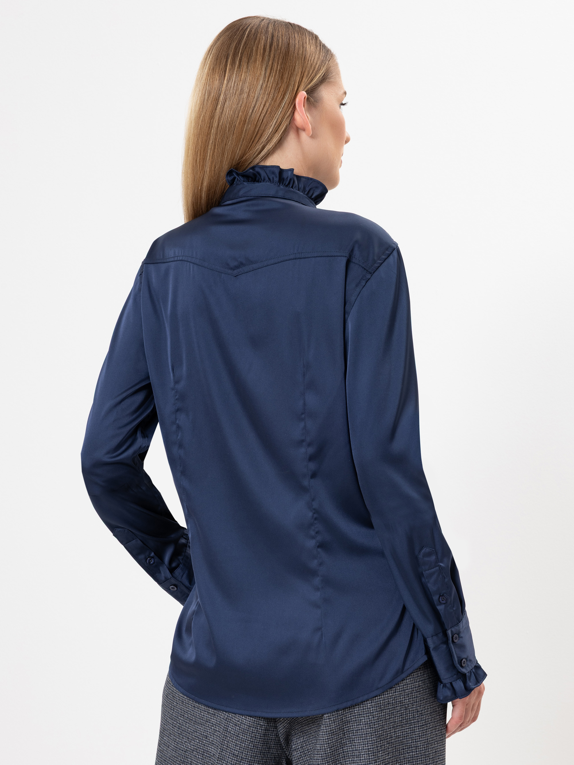 Eine Frau mit glattem, hellbraunem Haar ist von hinten zu sehen. Sie trägt eine langärmelige marineblaue Bluse mit gerafftem Kragen und Manschetten sowie eine graue Hose vor einem schlichten weißen Hintergrund.
